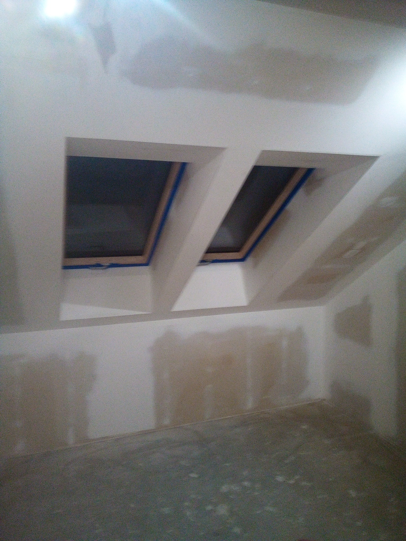 Pokój na poddaszu w trakcie prac wykończeniowych: dwa okna dachowe zabezpieczone taśmą malarską, ściany z widocznymi śladami szpachlowania, przygotowane do malowania.