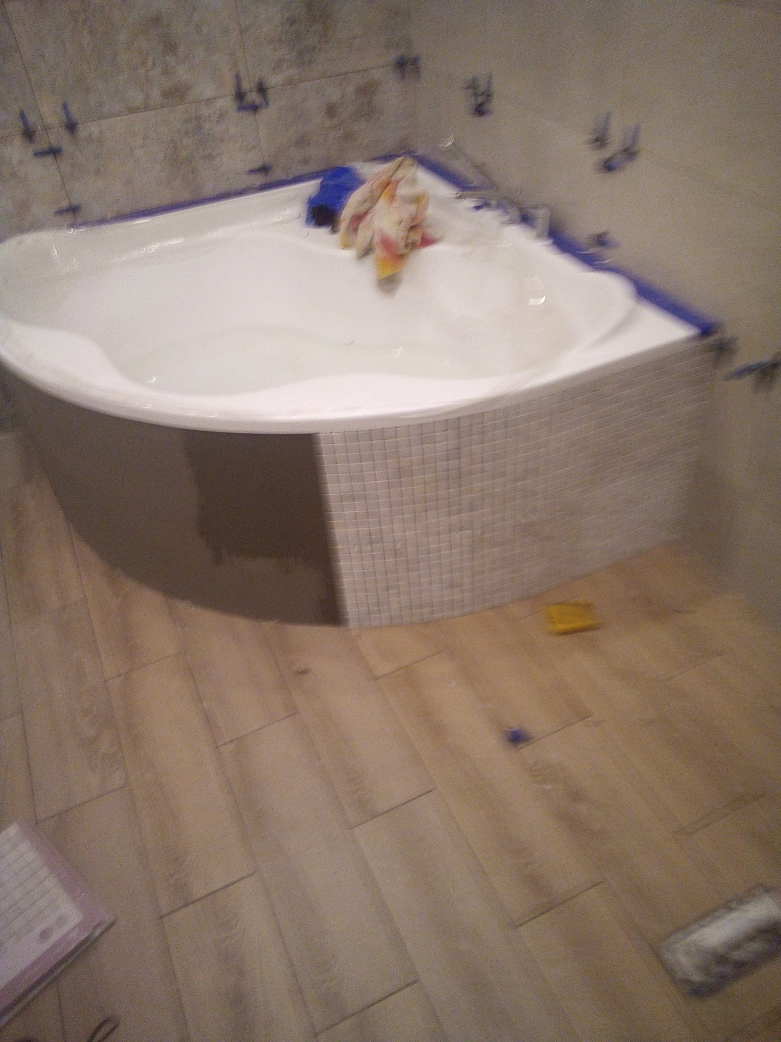 Narożna wanna obudowana mozaiką w trakcie montażu, widoczne krzyżyki dystansowe na ścianie i wzdłuż krawędzi wanny, drewnopodobna podłoga i akcesoria glazurnicze.