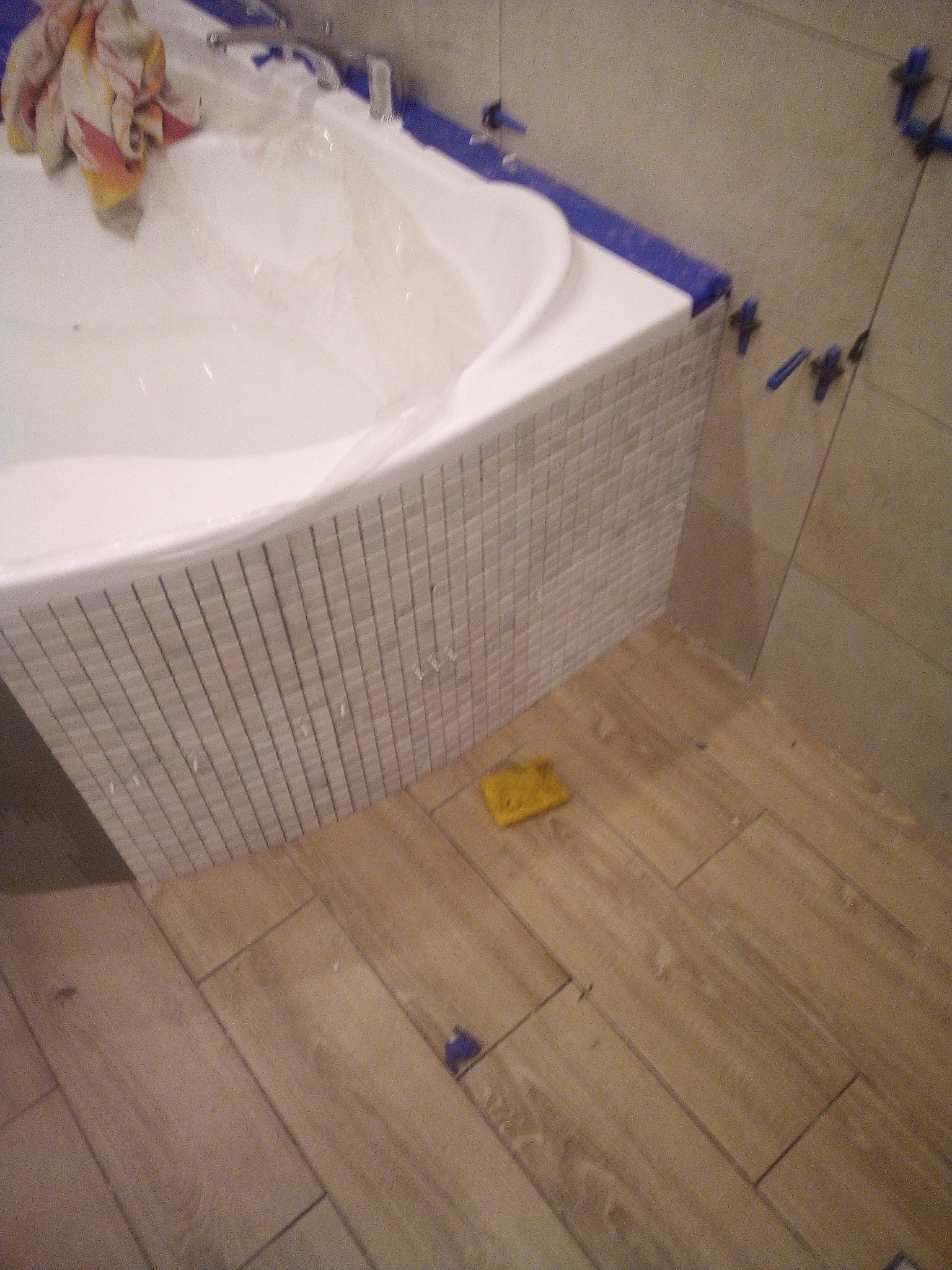 Mozaika z jasnych płytek na obudowie wanny w trakcie montażu, widoczne krzyżyki dystansowe, na wannie leży ręcznik, na podłodze płytki imitujące drewno oraz żółta gąbka.