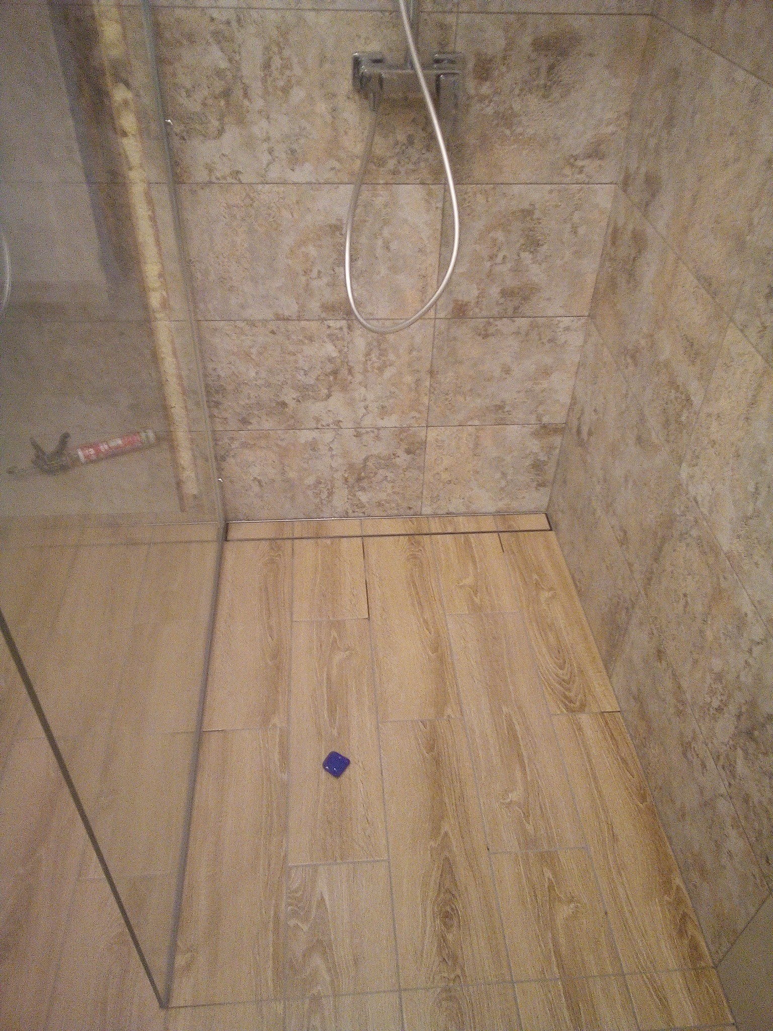 Kabina prysznicowa z odpływem liniowym w podłodze, wyłożona płytkami imitującymi drewno, z widoczną silikonową tubą za szybą i niebieskim elementem na podłodze.