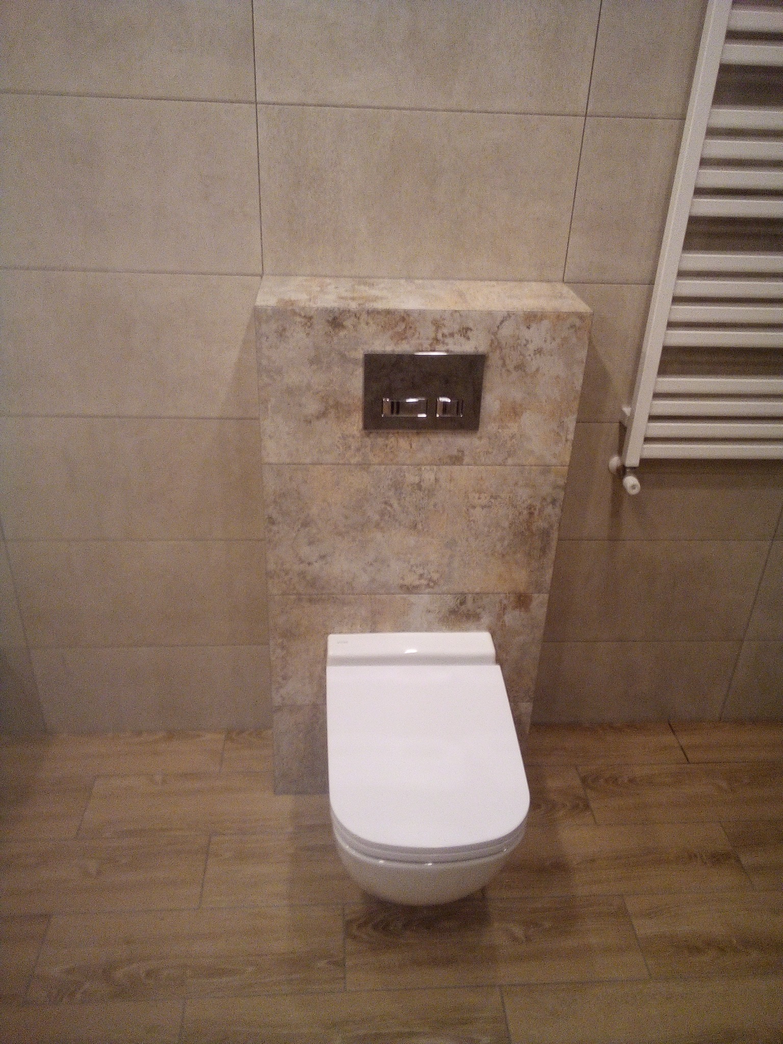 Wisząca toaleta z zabudową w płytkach imitujących kamień, chromowany panel spłukujący, grzejnik drabinkowy w łazience wykończonej płytkami w odcieniach beżu i brązu.