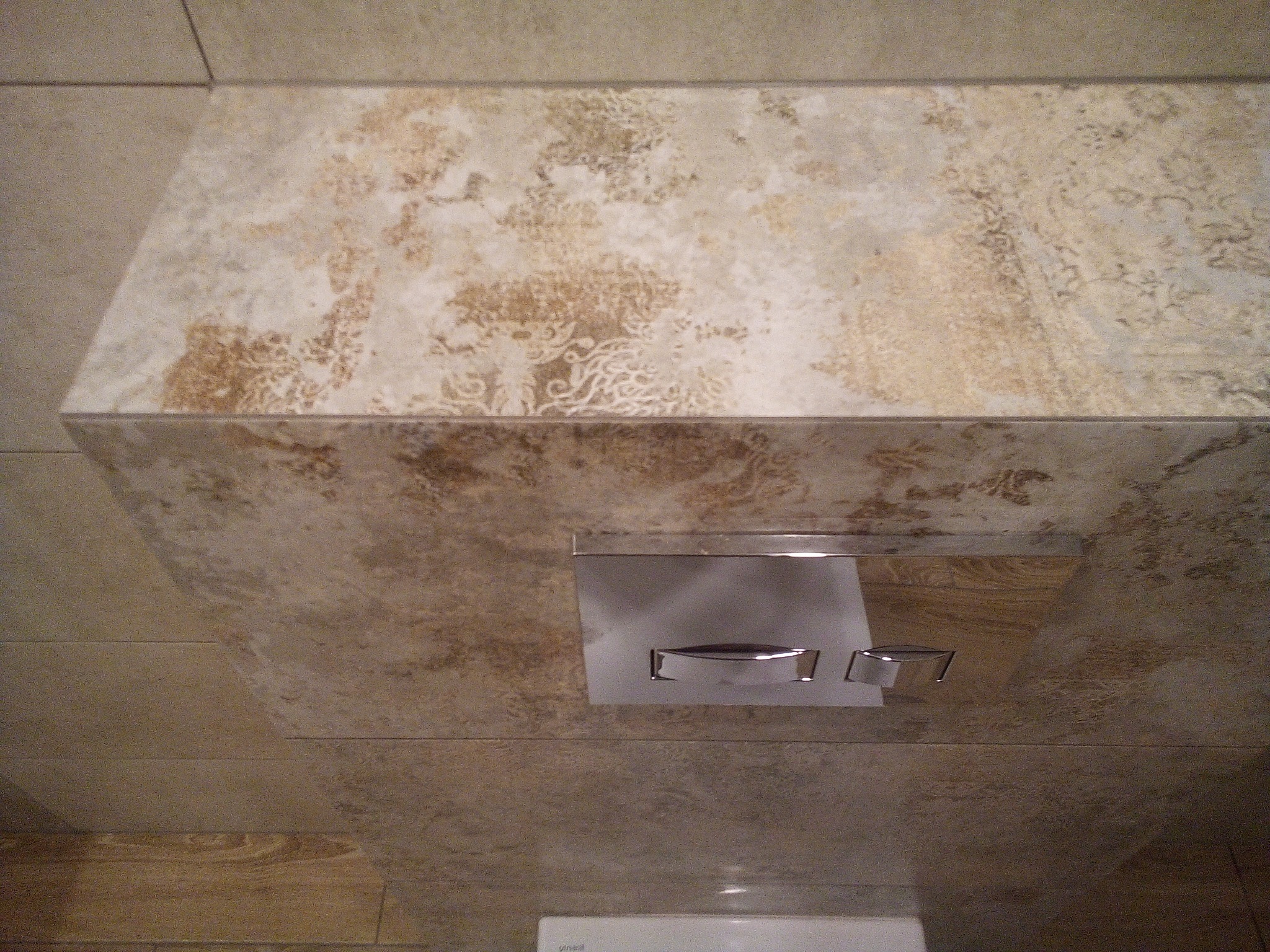 Ściana z beżowych, wzorzystych płytek ceramicznych w łazience, widoczny chromowany przycisk spłuczki podtynkowej.