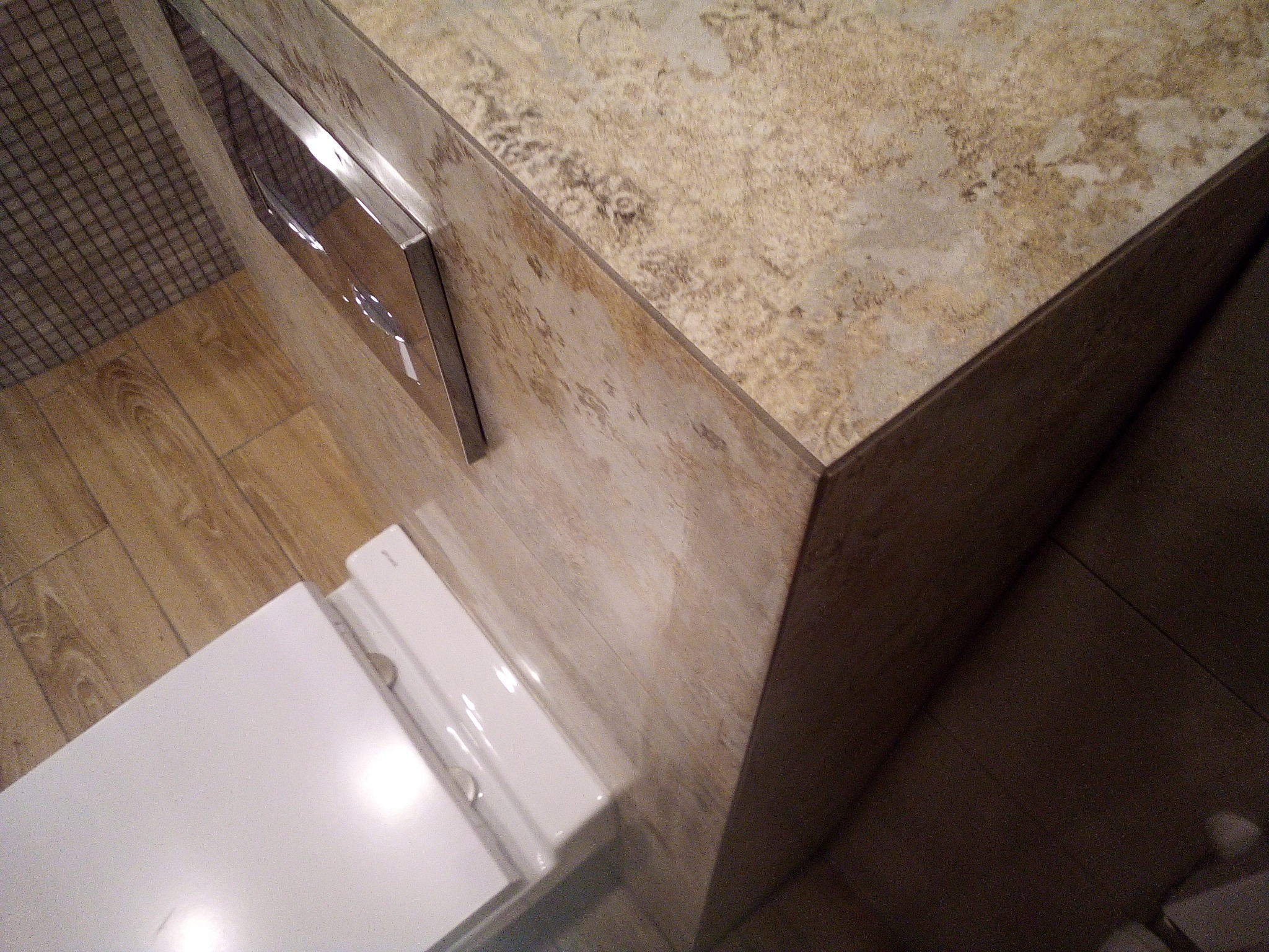 Fragment toalety z zabudowanym stelażem WC, wykończonej płytkami imitującymi kamień, z widocznym przyciskiem spłukującym ze stali nierdzewnej i drewnopodobną podłogą.