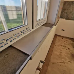 Montaż parapetu z szarej płytki przy oknie PCV z widokiem na zewnątrz, w trakcie prac wykończeniowych z widocznymi elementami instalacji i przygotowaniem pod glazurę na ścianie.