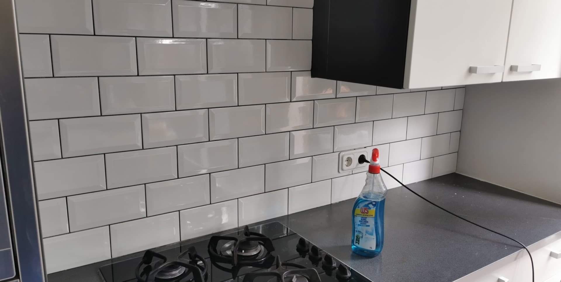 Kuchnia po remoncie: białe, prostokątne płytki ułożone na ścianie nad czarnym blatem i płytą gazową, z widocznym gniazdkiem elektrycznym i butelką płynu do czyszczenia.