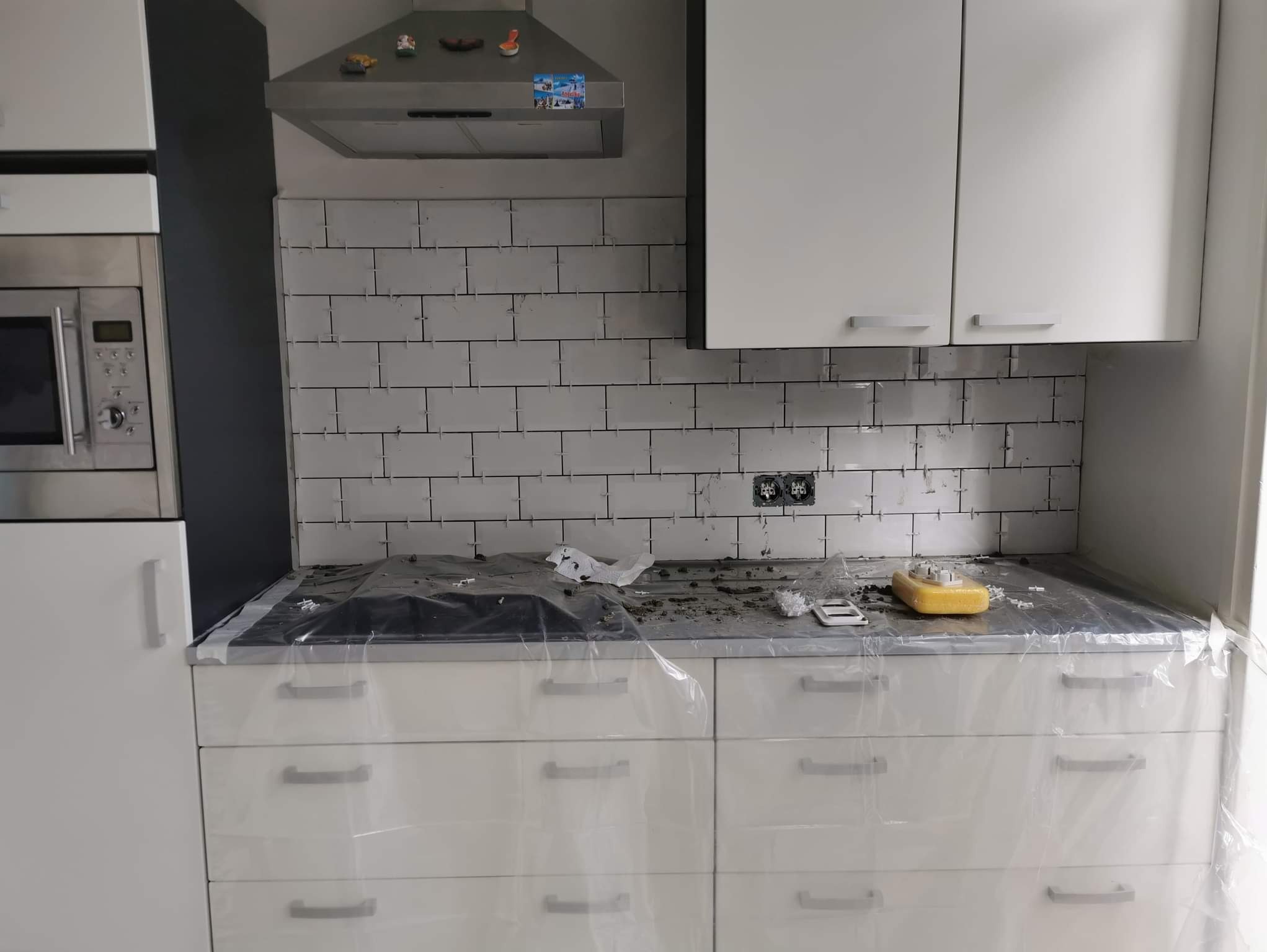 Białe płytki ułożone na ścianie w kuchni, z plastikowymi krzyżykami dystansowymi w spoinach, blat kuchenny zakryty folią ochronną, widoczne gniazdka elektryczne.