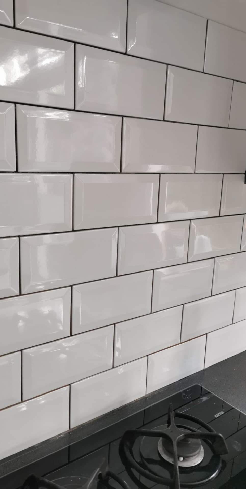 Białe, prostokątne płytki ułożone w cegiełkę na ścianie nad czarną płytą gazową. Widoczne fugi w kolorze ciemnoszarym.