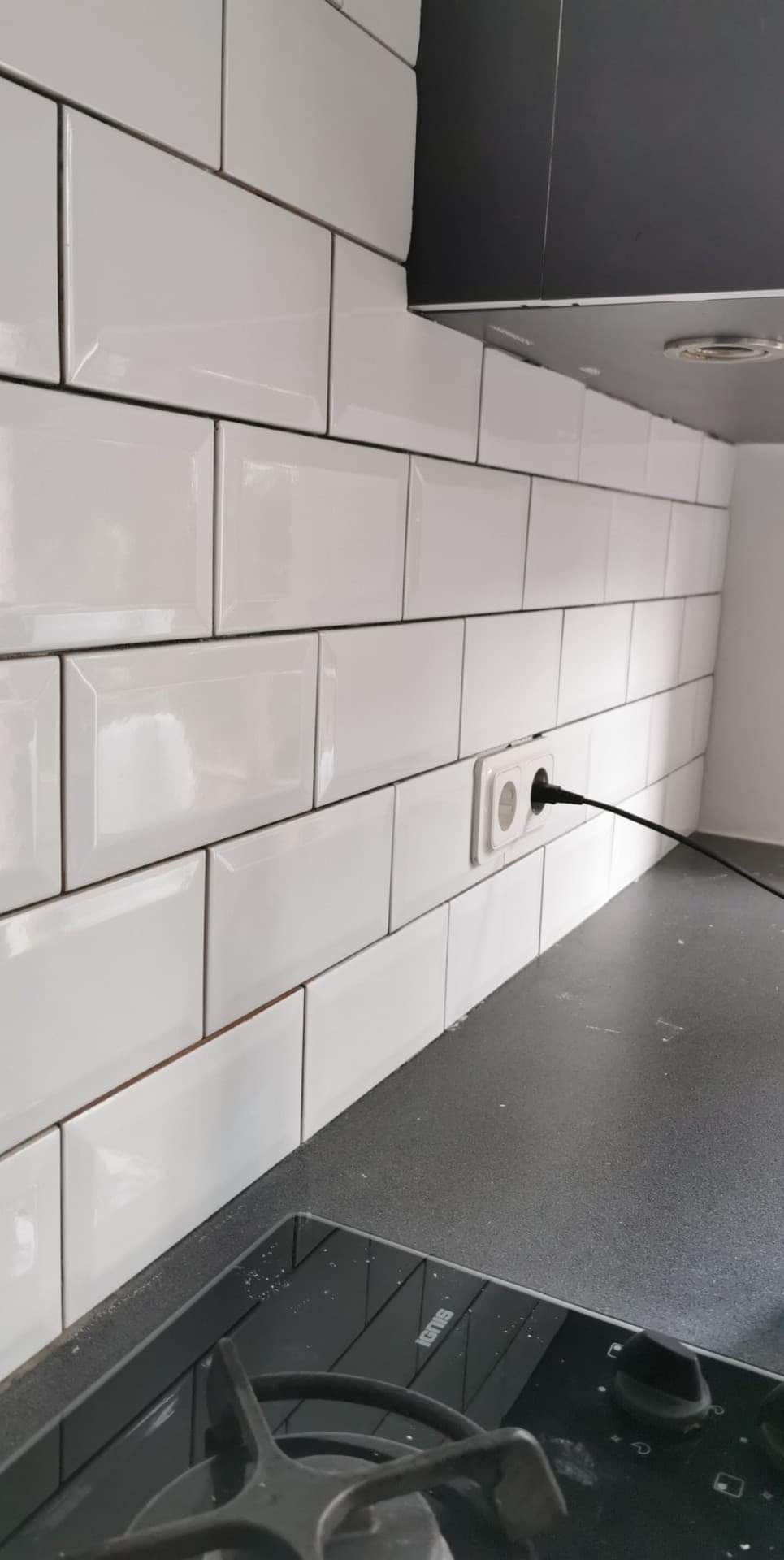 Białe, prostokątne płytki na ścianie nad blatem kuchennym, z widocznym gniazdkiem elektrycznym i czarnym przewodem, fragment kuchenki gazowej z przodu.