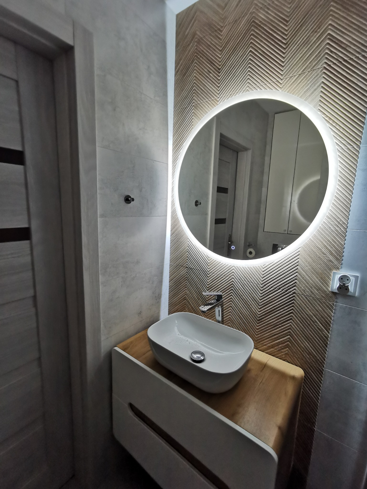 Nowoczesna łazienka z umywalką nablatową, okrągłym lustrem z podświetleniem LED i ścianą z dekoracyjną fakturą w jodełkę.