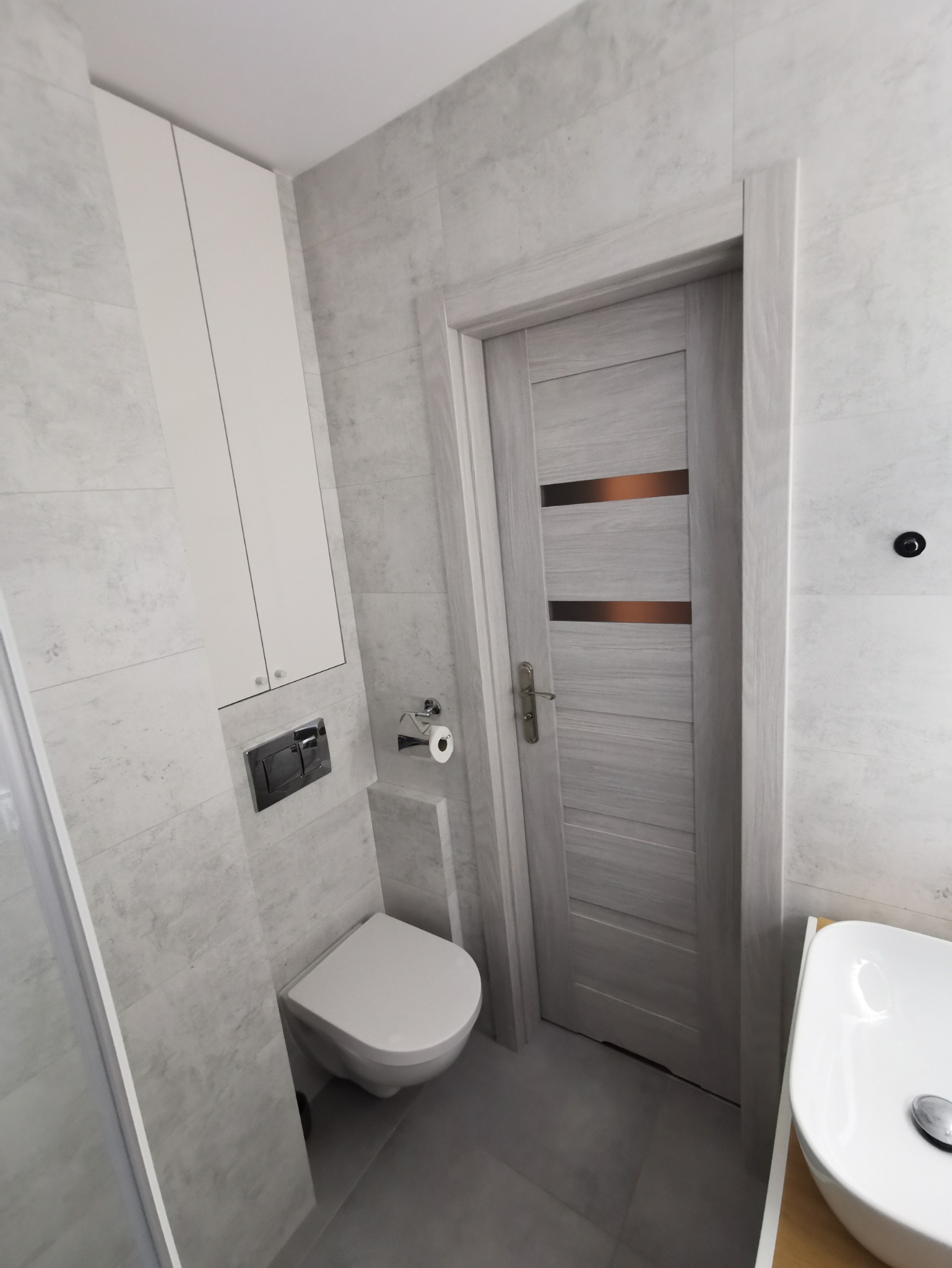 Nowoczesna łazienka w odcieniach szarości z podwieszaną toaletą, szafką wiszącą i drzwiami z poziomymi, metalicznymi wstawkami.