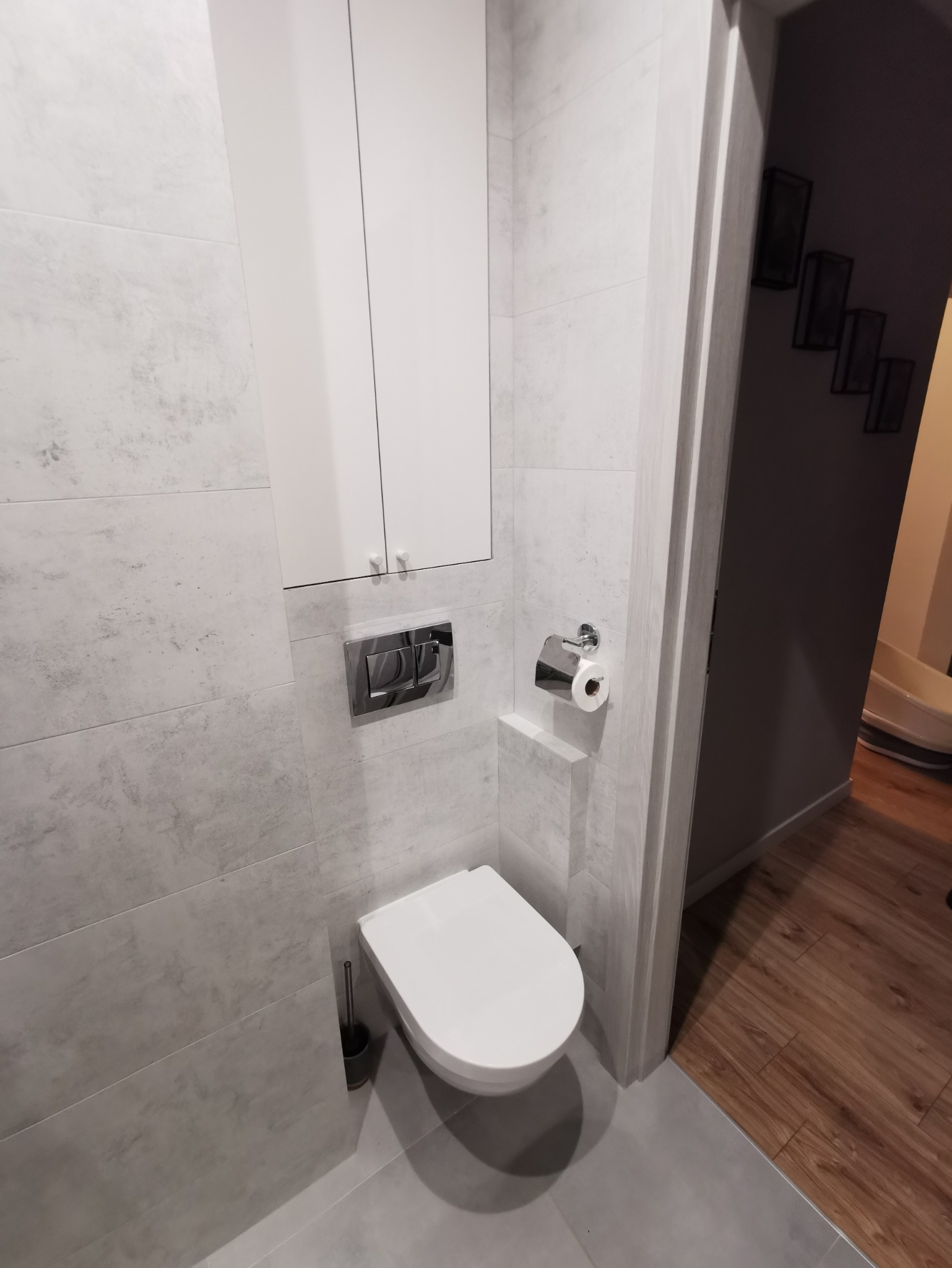 Nowoczesna toaleta w szaro-białej łazience z podwieszaną miską WC, szafką nad nią i chromowanym przyciskiem spłukującym, widok na drewnianą podłogę w sąsiednim pomieszczeniu.