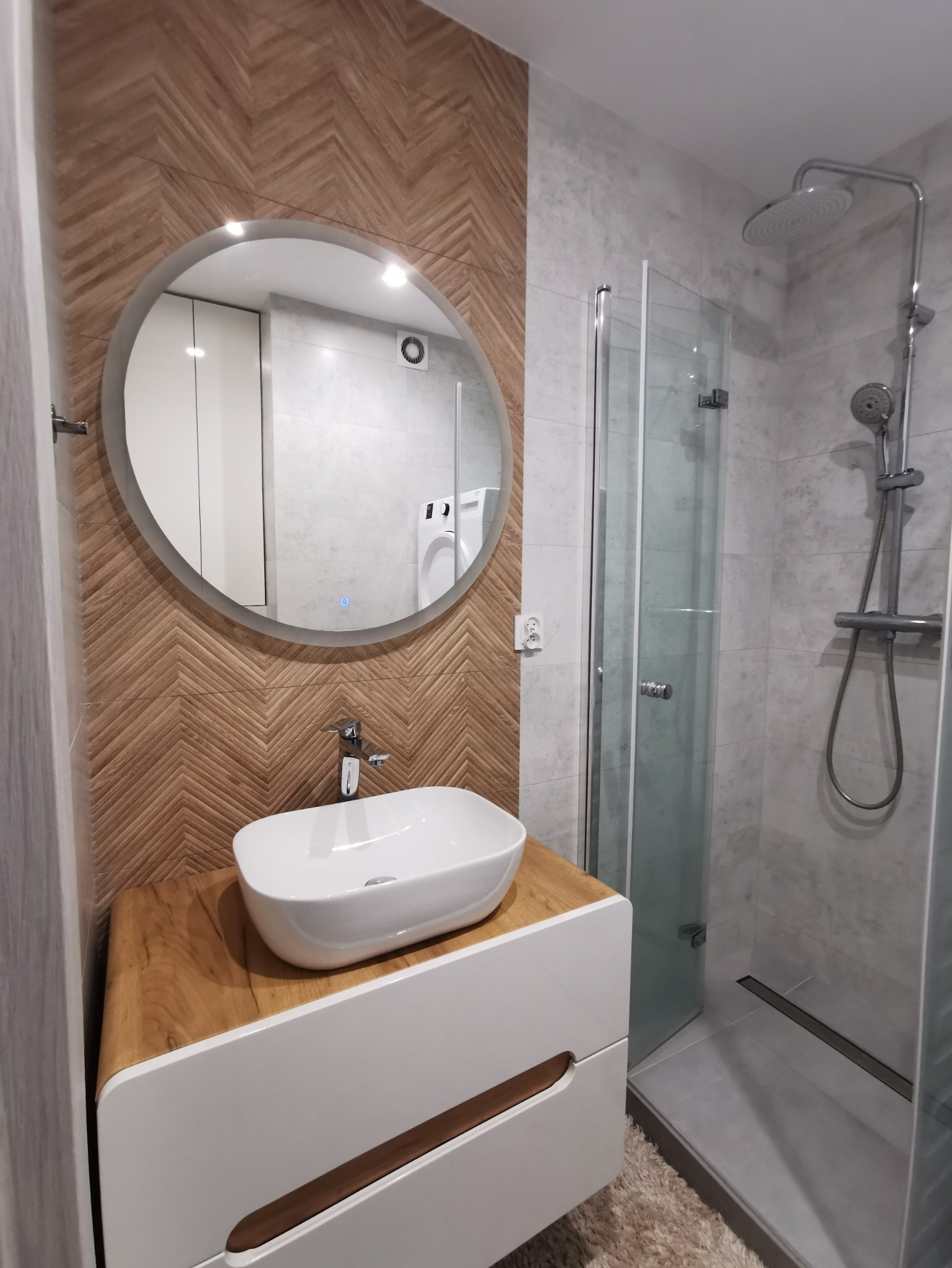 Nowoczesna łazienka z umywalką nablatową na szafce z drewnianym blatem, okrągłe lustro z podświetleniem LED, ściana z drewnopodobnych płytek ułożonych w jodełkę oraz kabina prysznicowa typu walk-in...