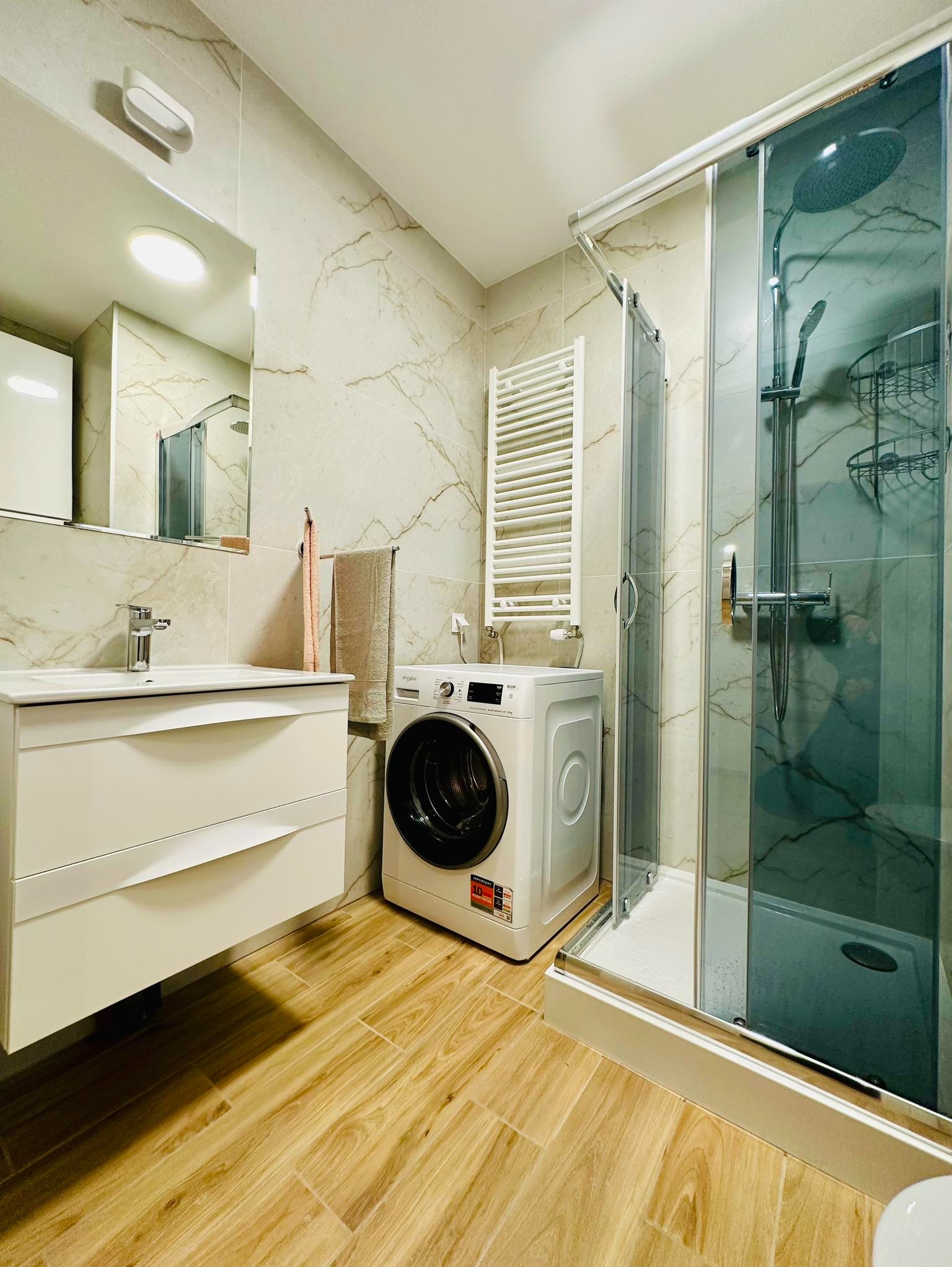 Jasna łazienka po remoncie z kabiną prysznicową, umywalką z szafką, pralką i grzejnikiem drabinkowym, wykończona płytkami imitującymi marmur i drewnem na podłodze.