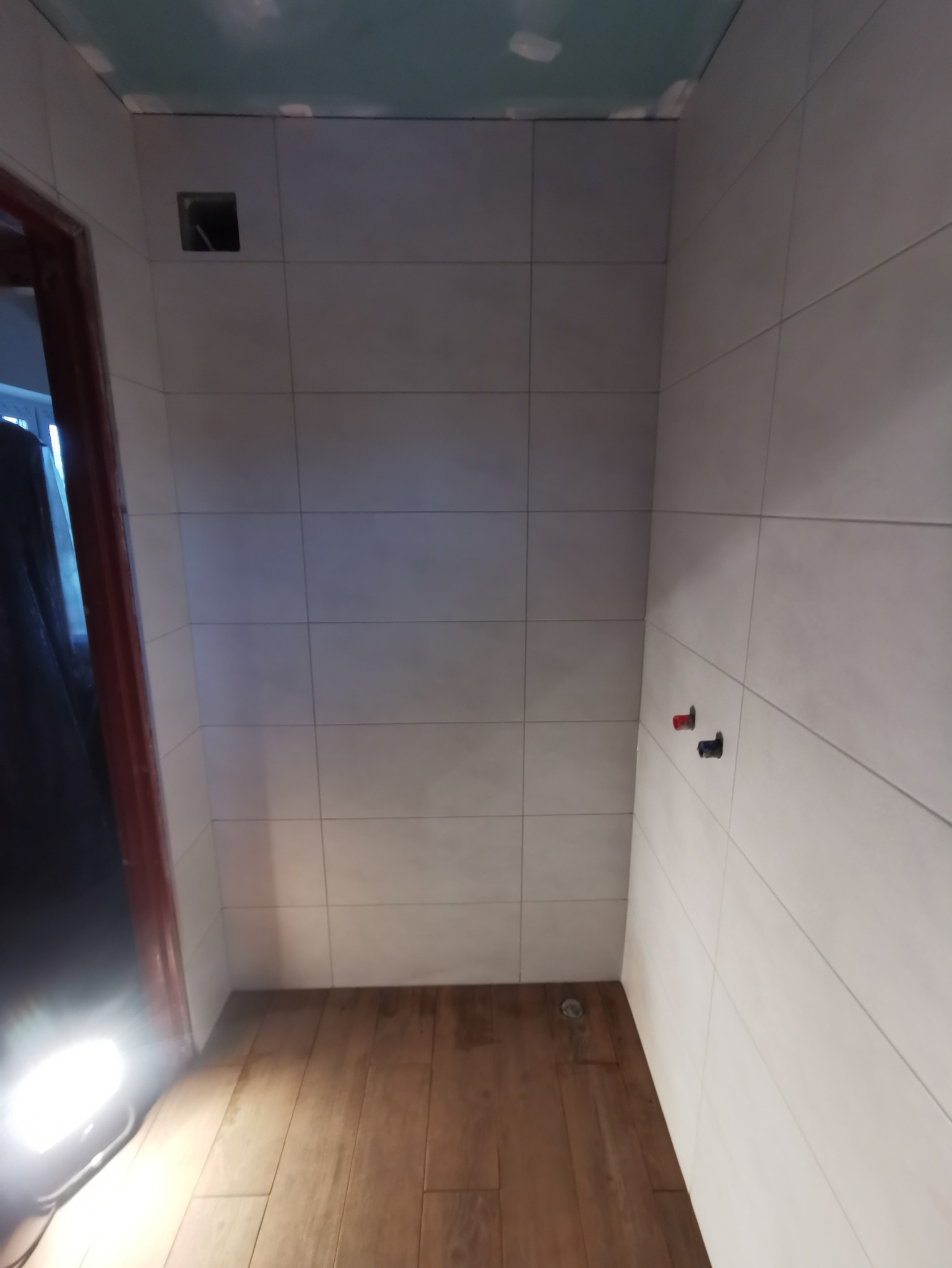 Wnętrze łazienki w trakcie prac wykończeniowych, widoczne szare płytki na ścianach, drewnopodobna podłoga, otwór wentylacyjny w górnej części ściany i wystające przyłącza wodne.