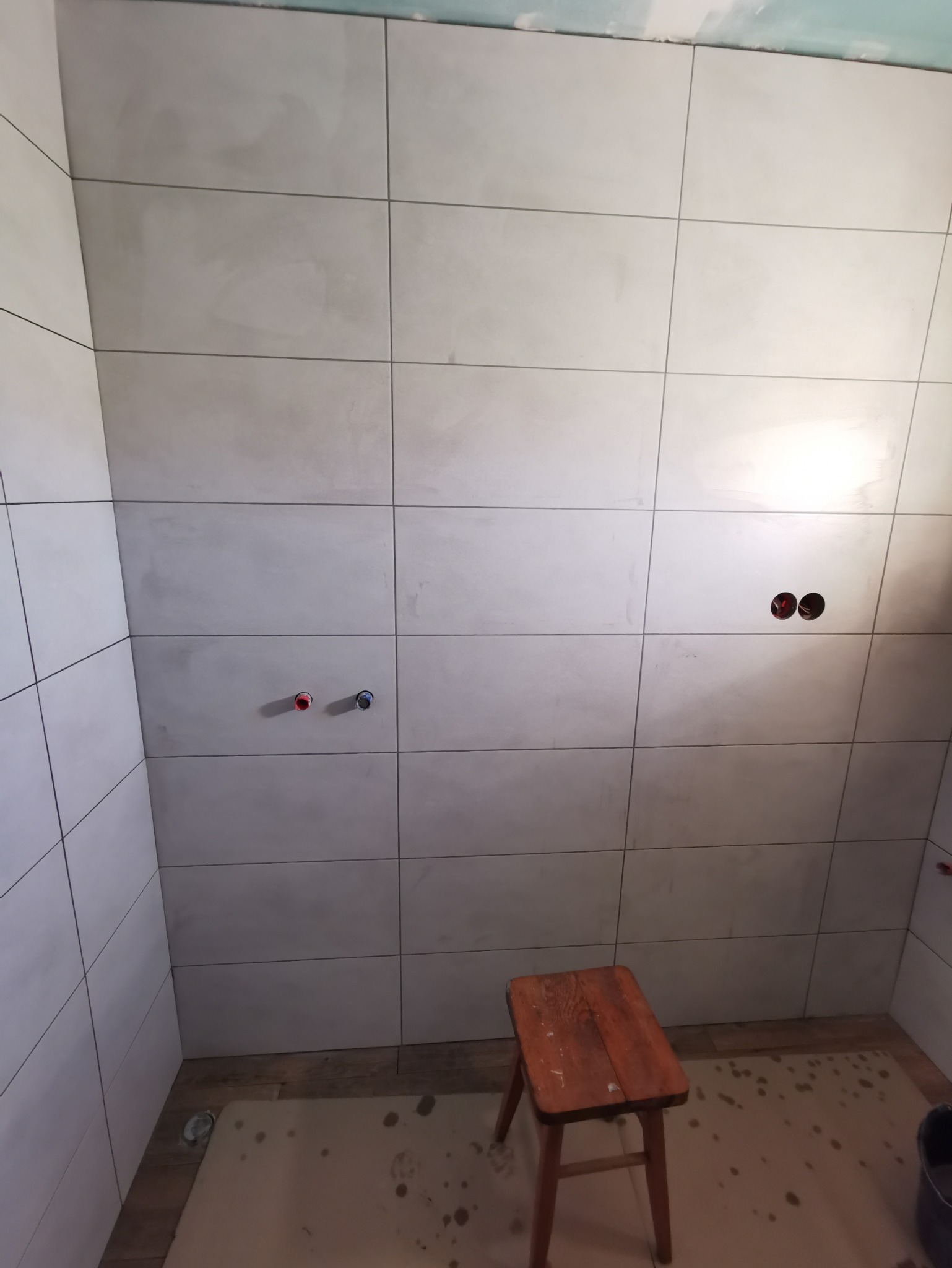 Ściany łazienki wyłożone jasnymi, matowymi płytkami ceramicznymi w trakcie prac wykończeniowych. Widoczne przygotowane otwory na baterie i drewniany stołek na podłodze zabezpieczonej kartonem.