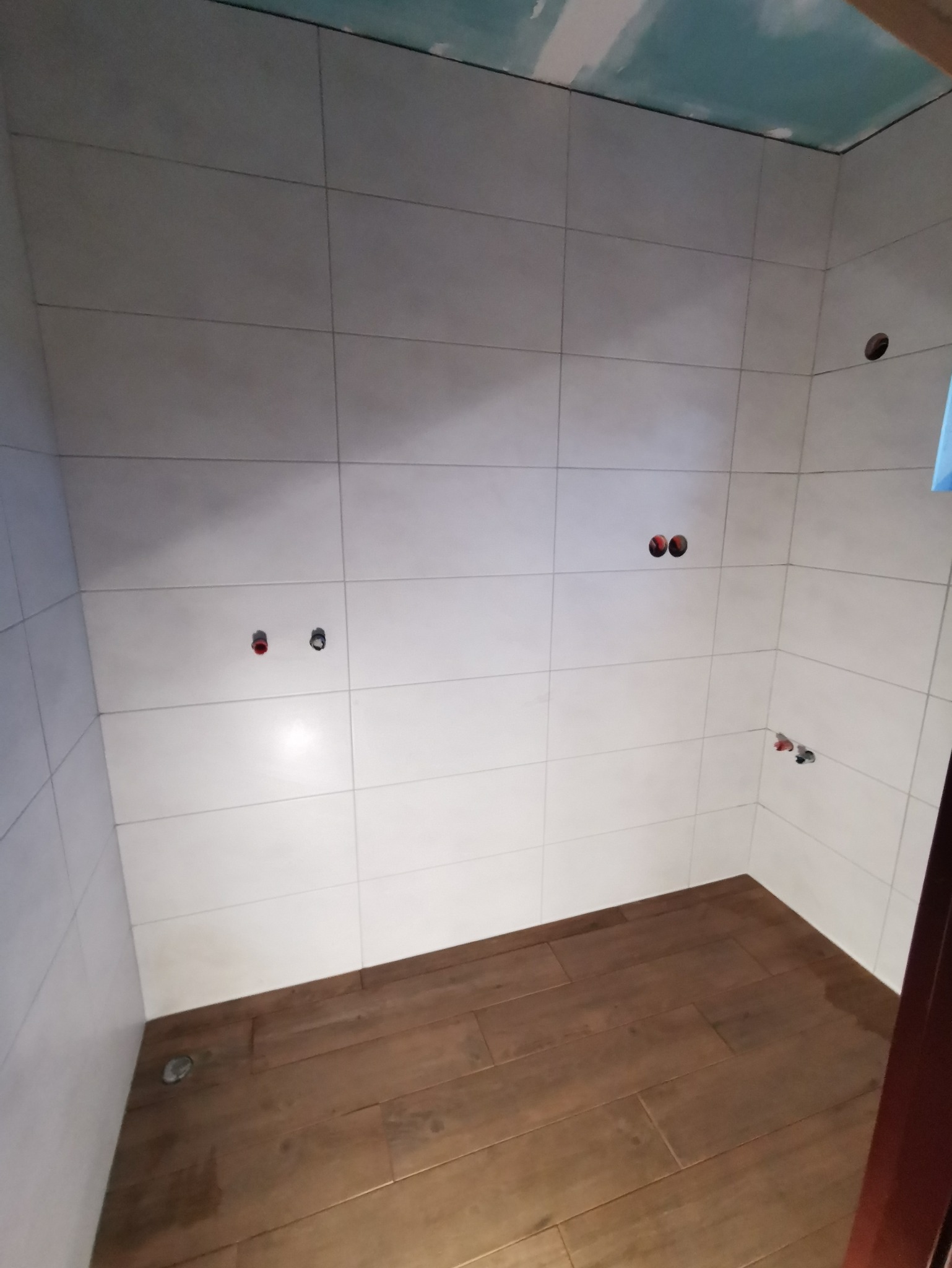 Wykończona łazienka z białymi płytkami ściennymi i drewnopodobną podłogą, widoczne otwory na armaturę i niedokończony sufit z płyt gipsowo-kartonowych.