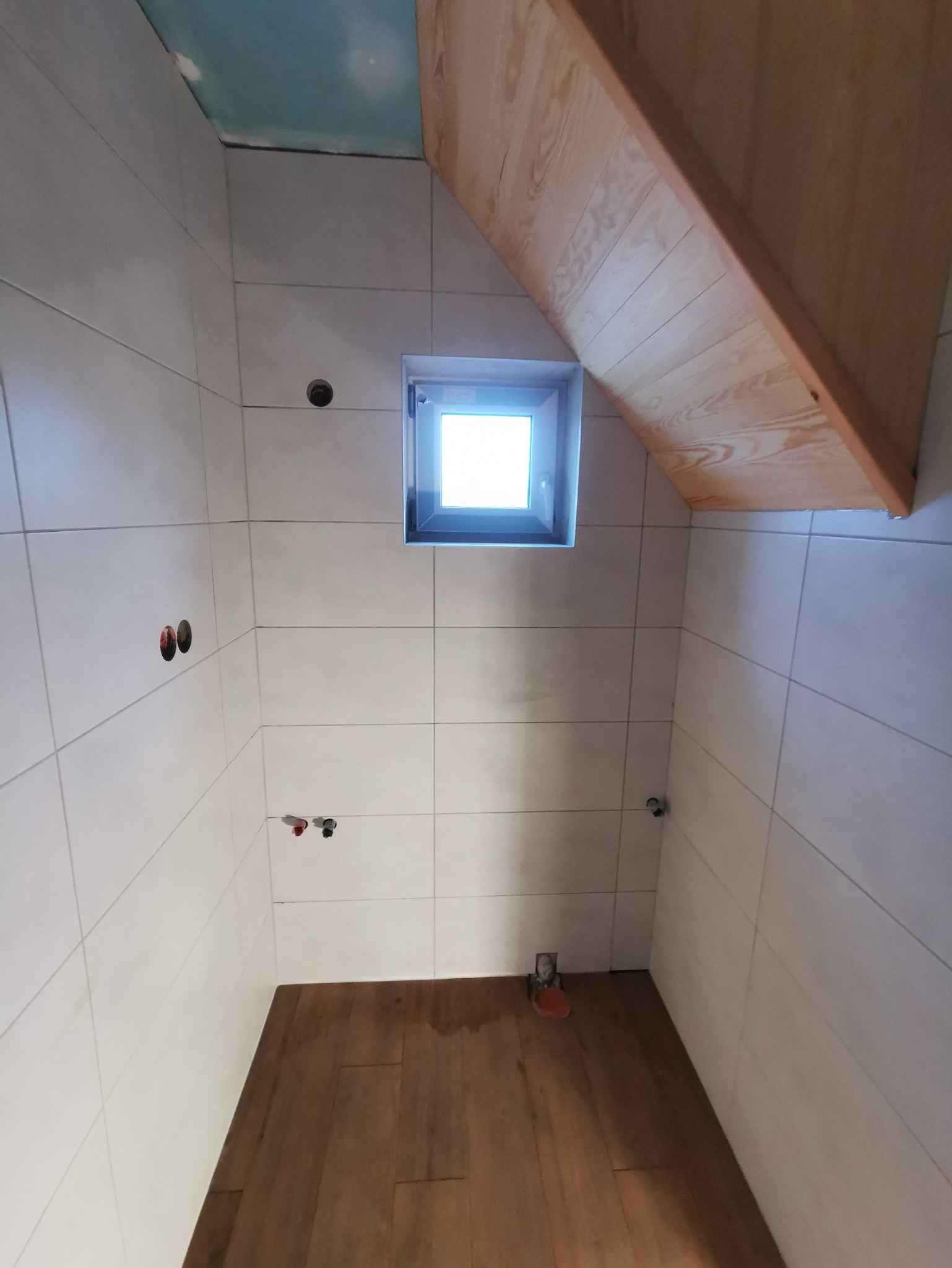 Wnętrze łazienki w trakcie wykańczania, z widocznymi białymi płytkami na ścianach, drewnianą podłogą, oknem i drewnianym elementem konstrukcyjnym pod skosem sufitu.