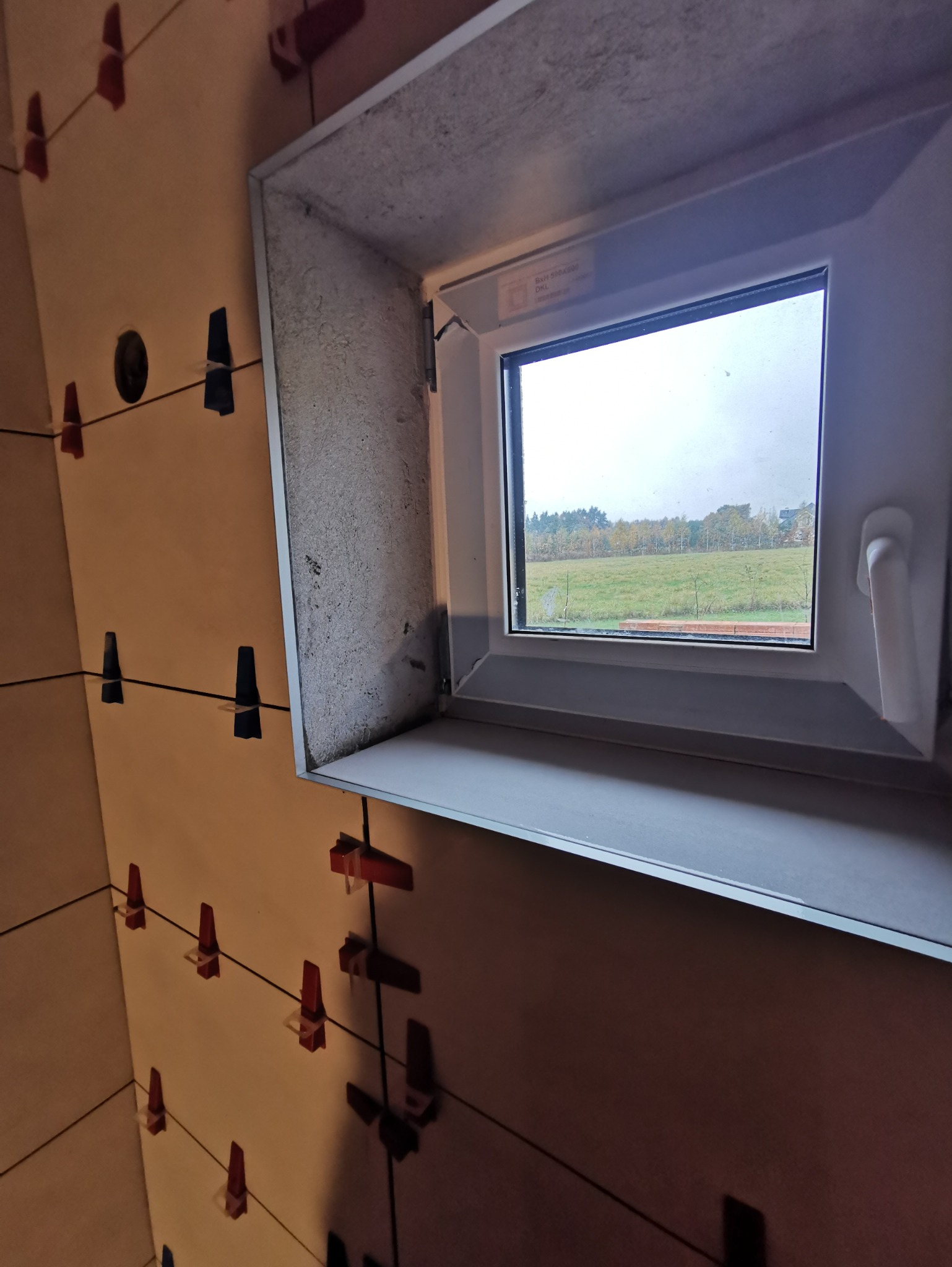 Ściana z jasnych płytek, w trakcie układania, z widocznymi klipsami poziomującymi i oknem z widokiem na zielone pole.