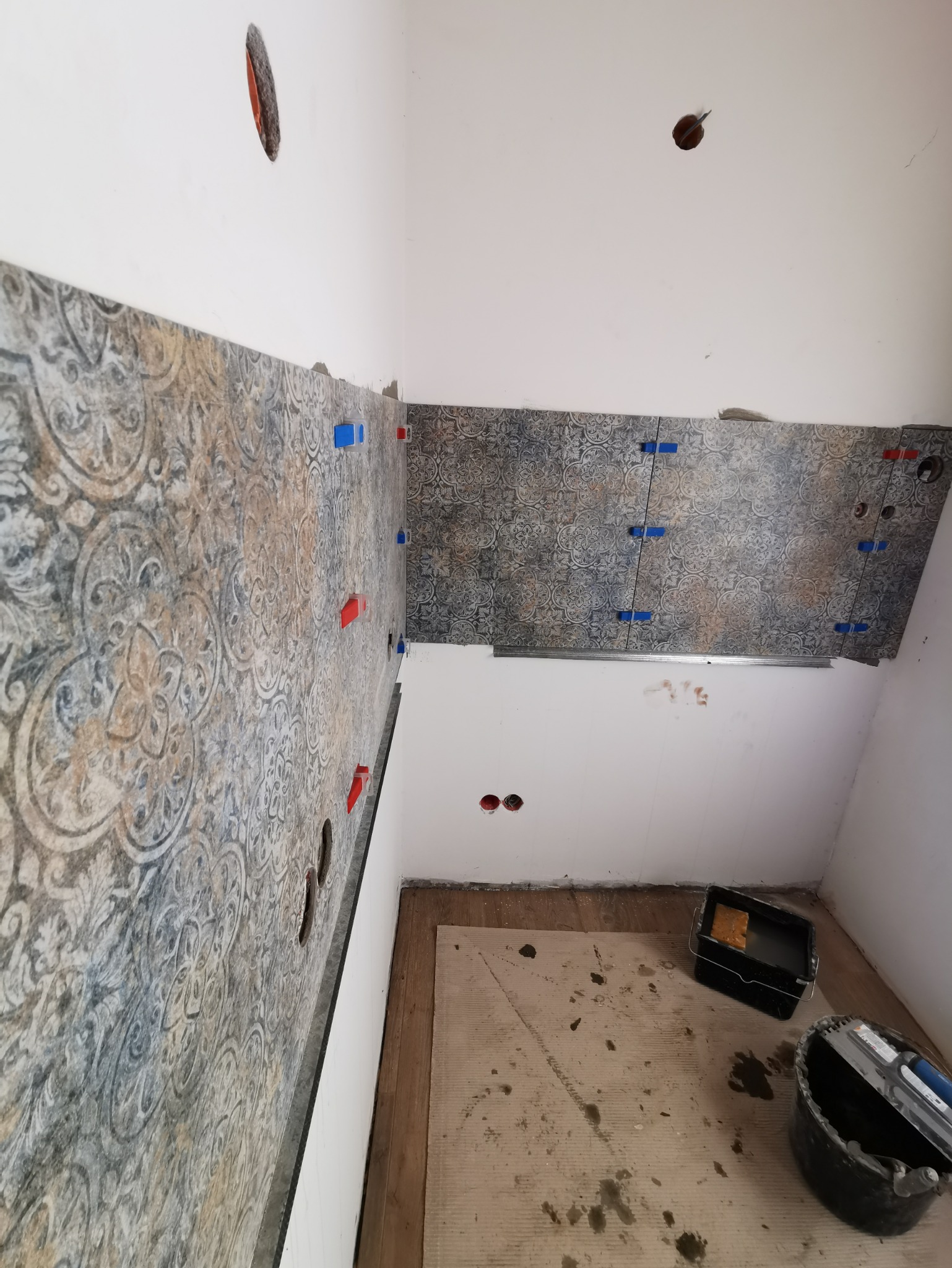 Ściany w trakcie układania dekoracyjnych płytek z użyciem niebieskich i czerwonych krzyżyków dystansowych. Na podłodze karton ochronny, wiadro i szpachelka do kleju.