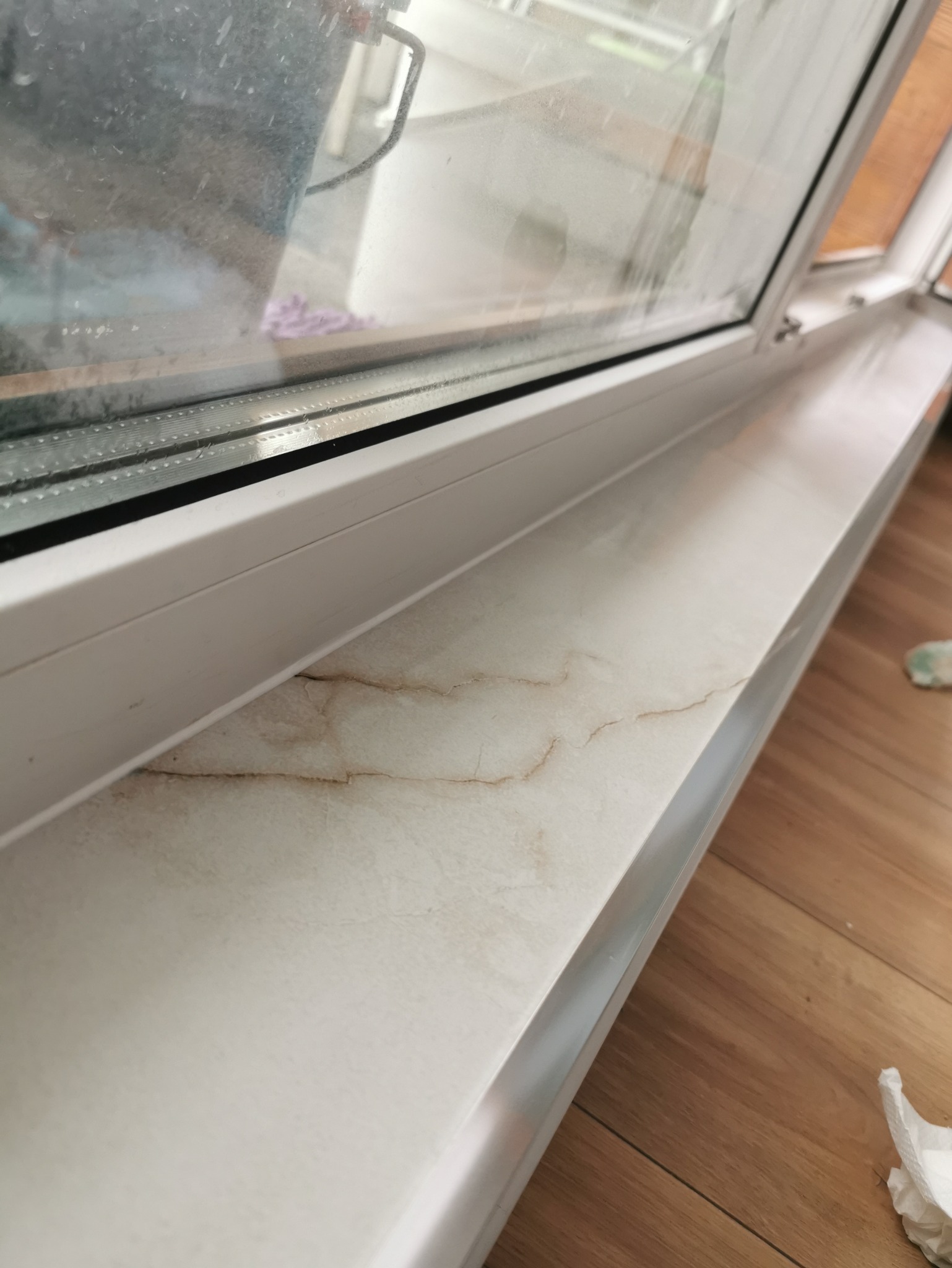 Nowy, biały parapet okienny z wzorem imitującym marmur, zamontowany pod brudnym oknem, widoczne drewniane panele podłogowe.