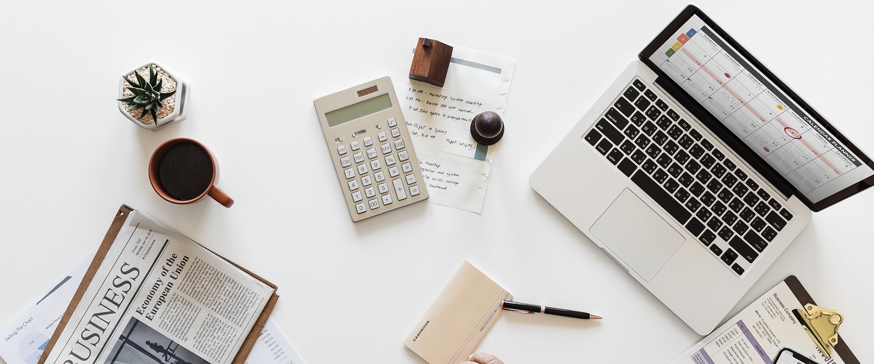 Widok z góry na biurko z laptopem z kalendarzem, kalkulatorem, notatnikiem, długopisem i gazetą biznesową. Stonowana kolorystyka, minimalistyczny styl.