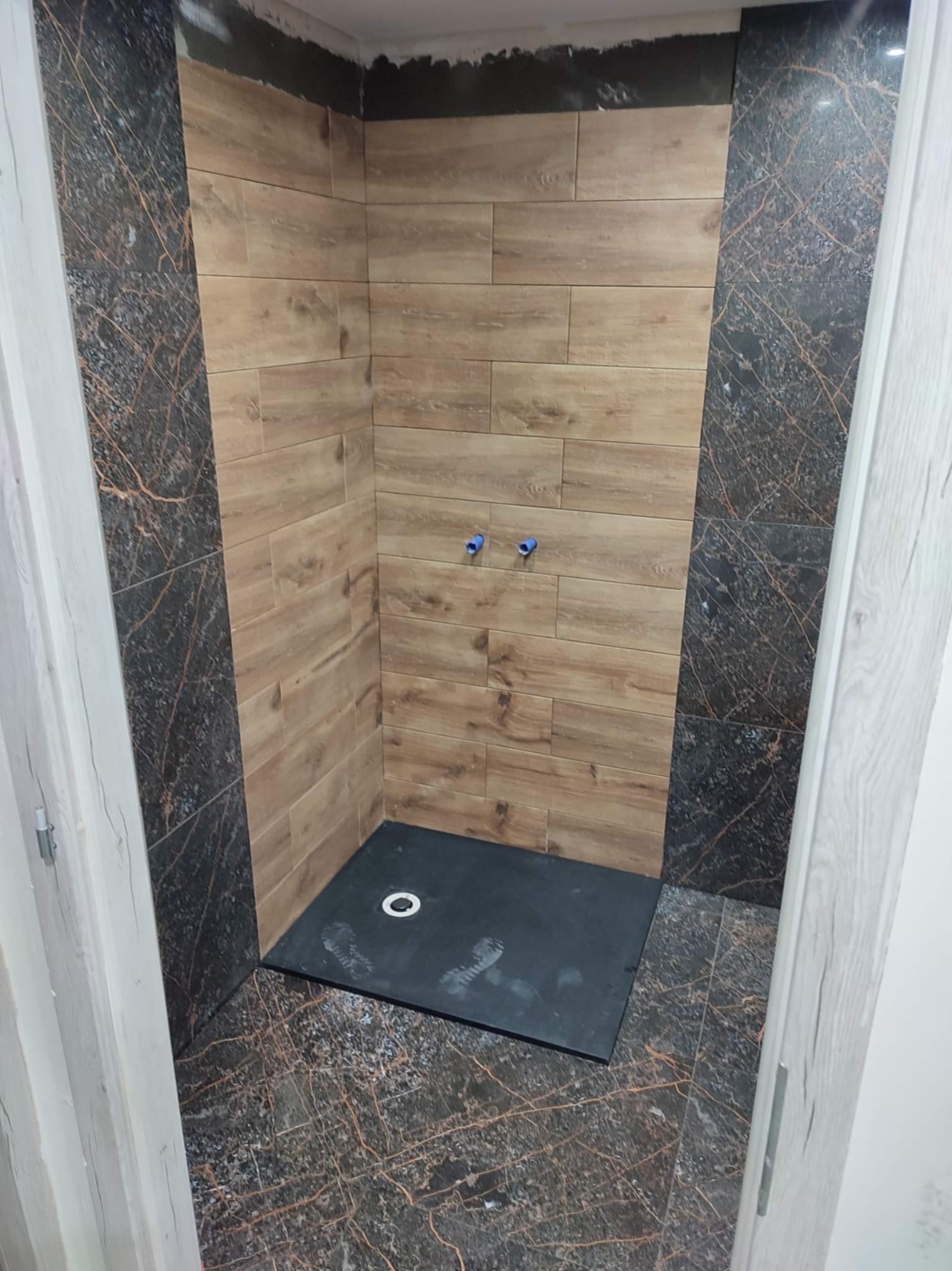 Niedokończona kabina prysznicowa z brązowymi płytkami imitującymi drewno na ścianach i ciemnymi płytkami z marmurkowym wzorem na podłodze oraz pionowym pasie na ścianie, widoczne niebieskie rury...
