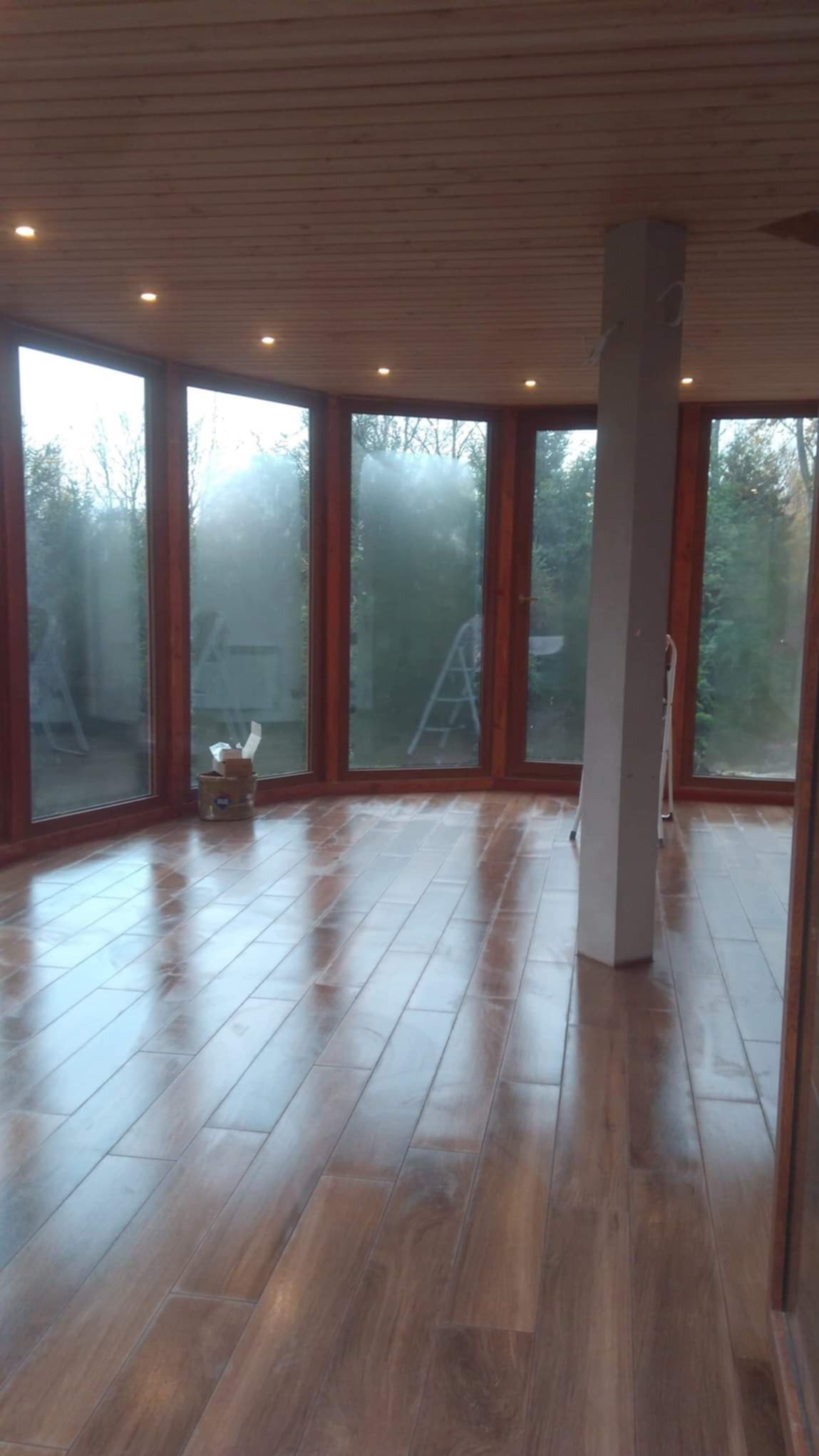 Wnętrze jasnej oranżerii z drewnianą podłogą imitującą panele, dużymi oknami z drewnianymi ramami, drewnianym sufitem z wbudowanymi punktowymi lampami i centralnym filarem. Częściowo widoczne...