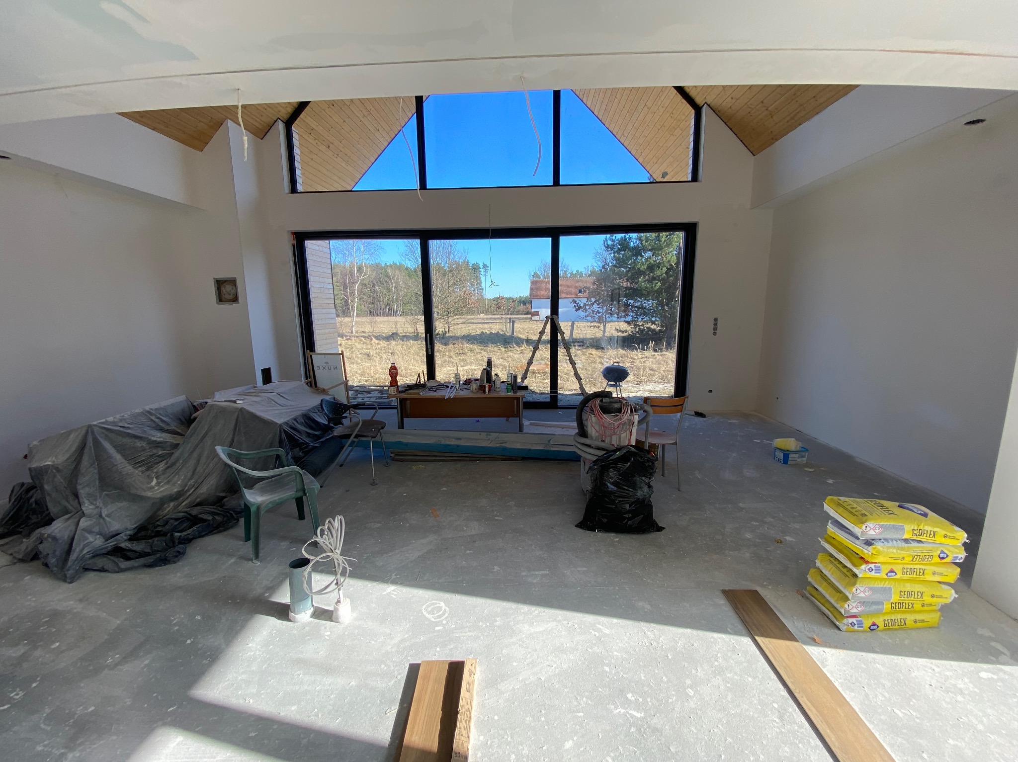 Wnętrze w trakcie prac wykończeniowych: surowy beton, worki z zaprawą, okna z widokiem na zewnątrz, drewniane elementy konstrukcyjne dachu. Widoczne narzędzia i materiały budowlane.