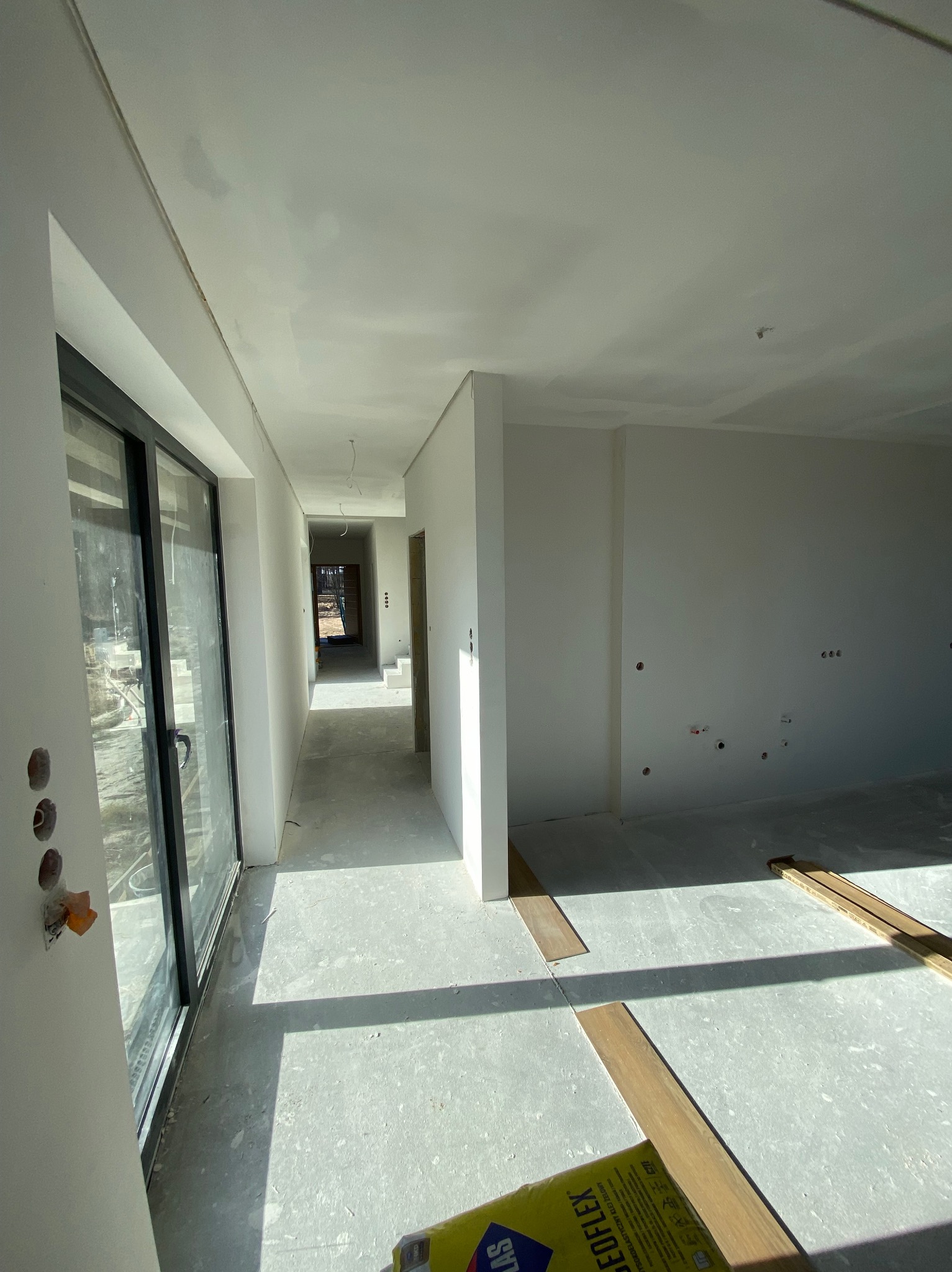 Wnętrze w trakcie wykończenia: gładkie, białe ściany, surowa podłoga, duże okno przesuwne. Widoczne przygotowania do montażu podłogi.