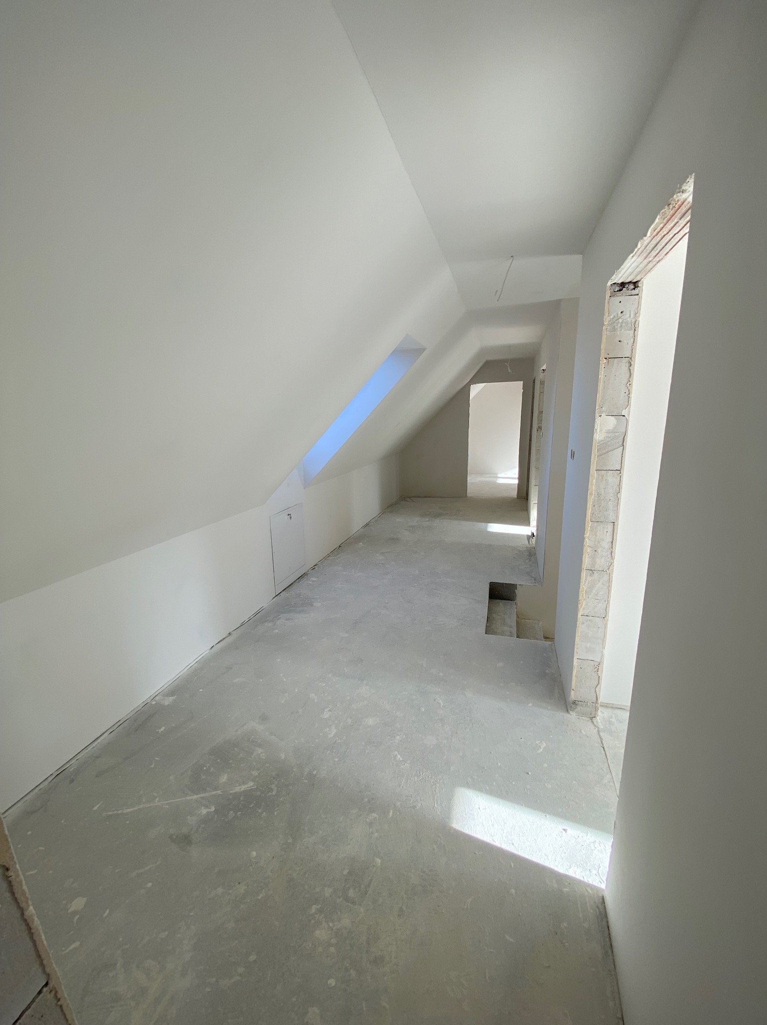 Białe, wykończone poddasze ze skosami i oknem dachowym. Widoczne surowe ściany i wylewka podłogowa. W tle widoczne kolejne pomieszczenia w trakcie prac.