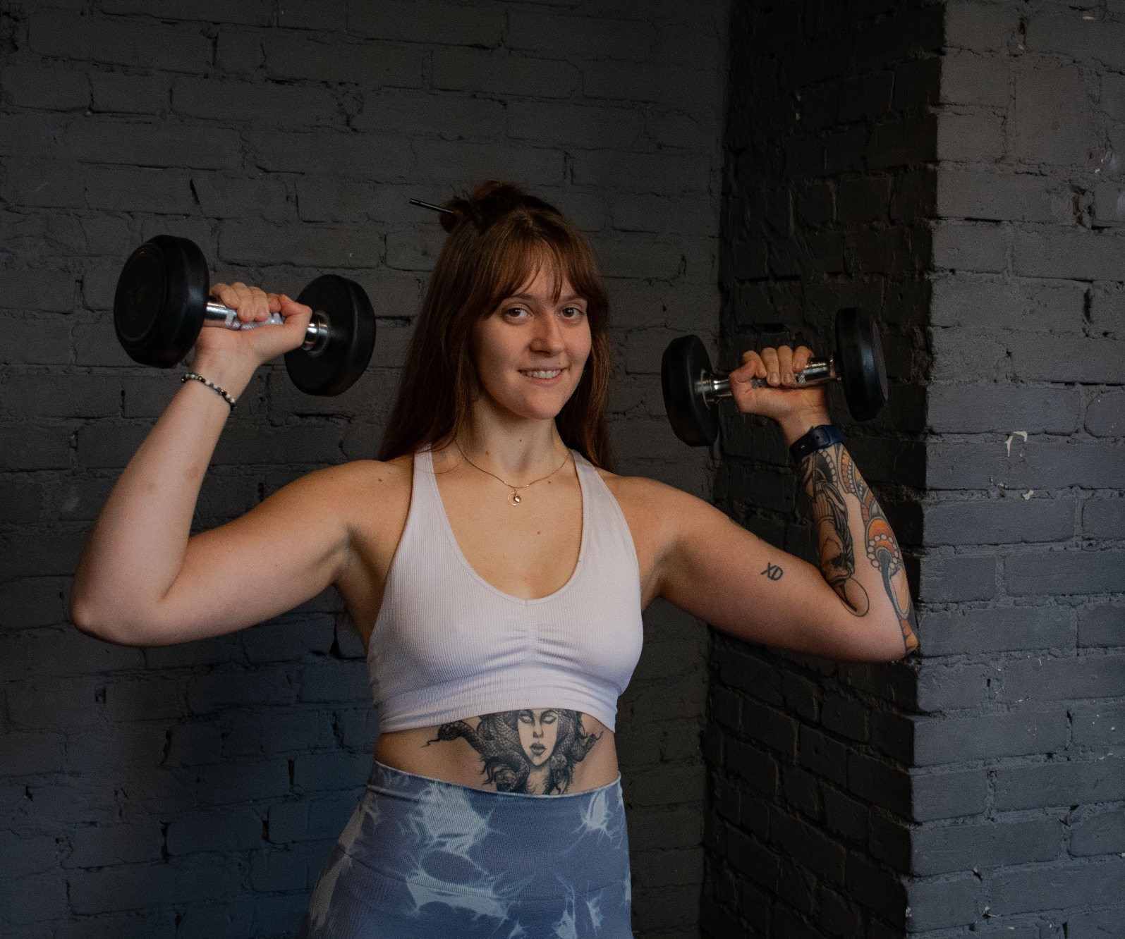 Uśmiechnięta kobieta w sportowym stroju, z widocznymi tatuażami na ramieniu i brzuchu, ćwiczy z hantlami na tle ciemnej, ceglanej ściany.