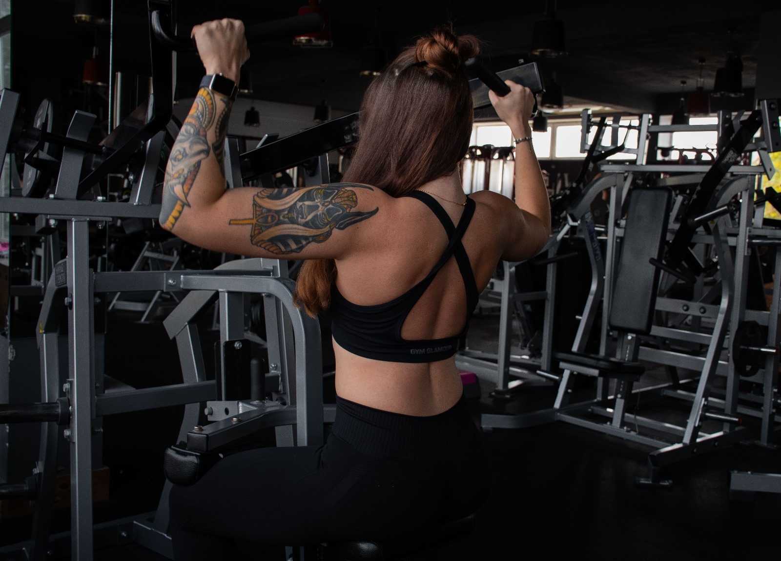 Kobieta z tatuażami na ramionach ćwiczy na maszynie w siłowni, widok z tyłu, włosy upięte w kok, ubrana w sportowy top i legginsy.