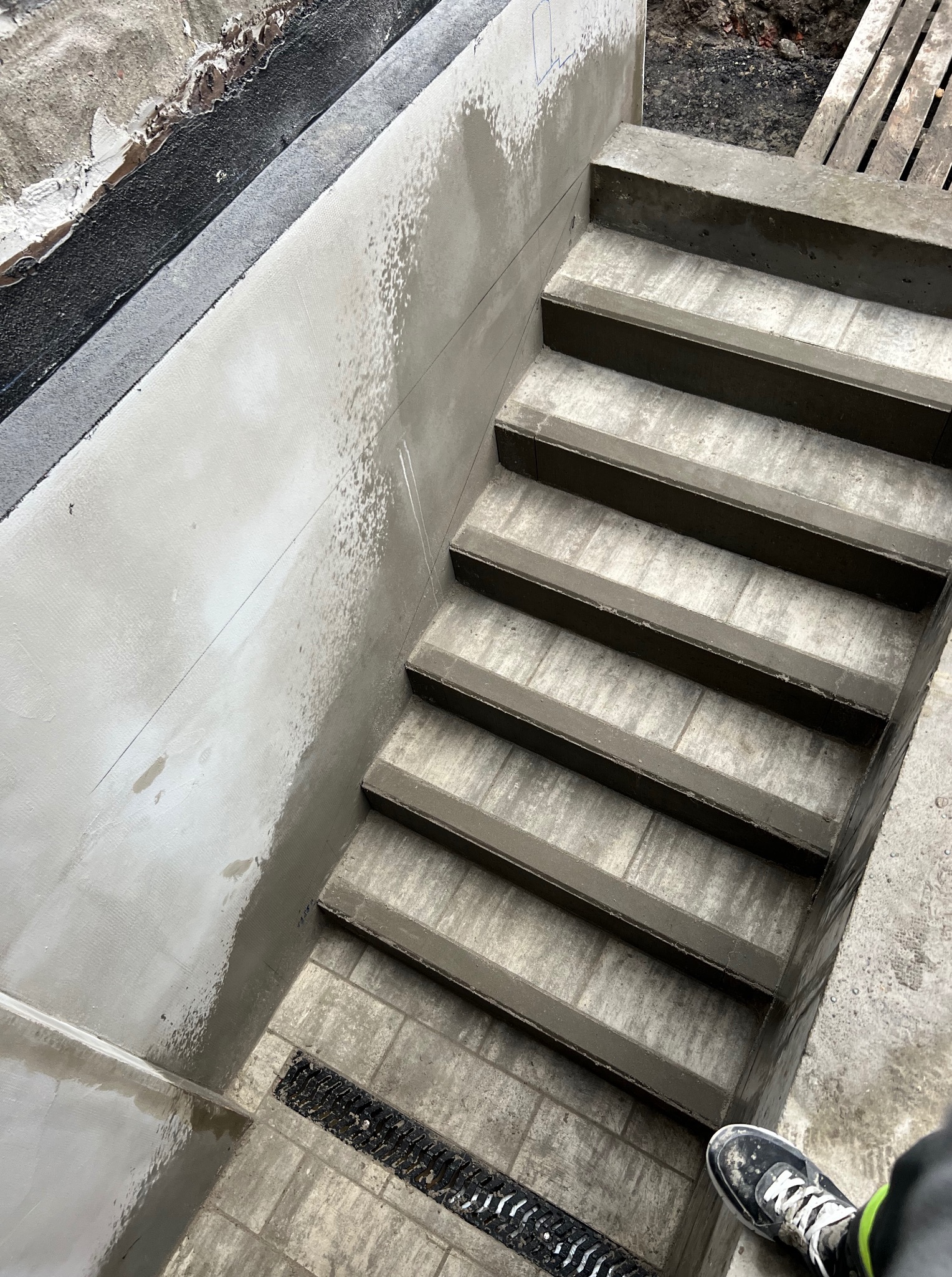 Betonowe schody zewnętrzne w trakcie budowy, widok z góry. Świeży beton na ścianach i stopniach, widoczny odpływ liniowy. But na dole kadru.