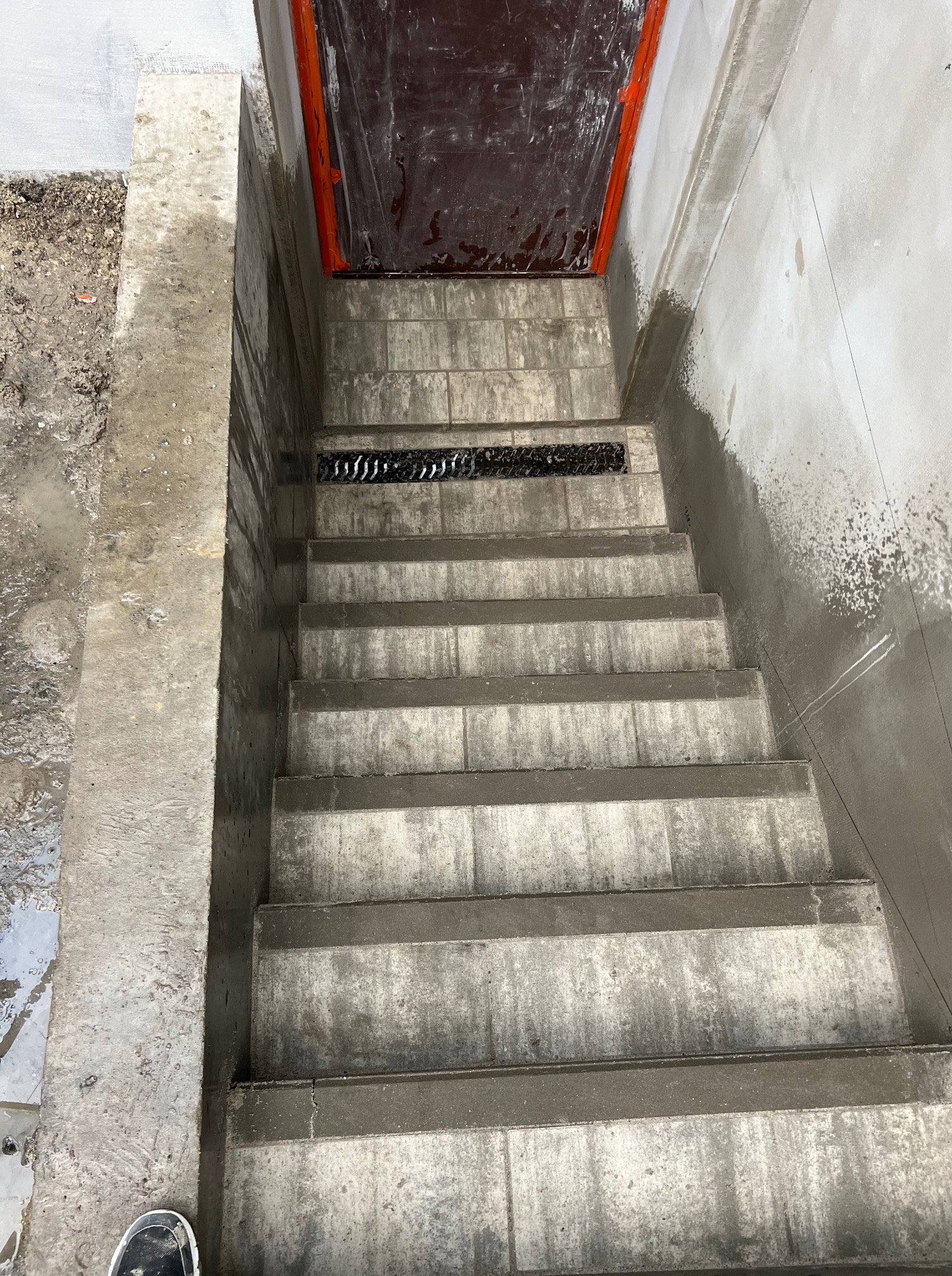 Betonowe schody z odpływem liniowym w łazience, widok z góry, surowy stan wykończenia, pomarańczowe obramowanie drzwi.