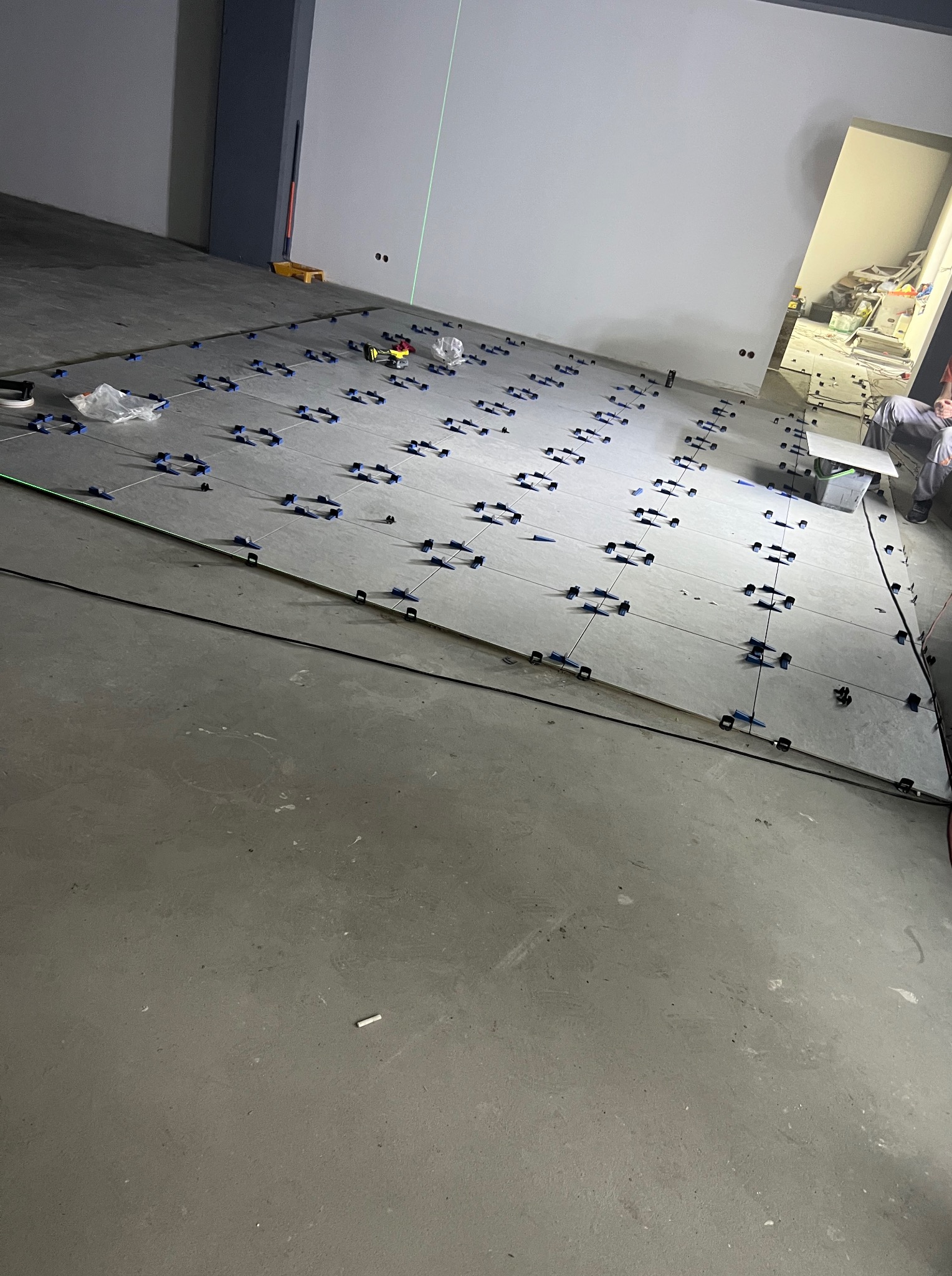 Układanie szarych płytek z użyciem klipsów poziomujących i lasera krzyżowego. Widoczny fragment osoby układającej płytki. Praca w toku, surowy stan.