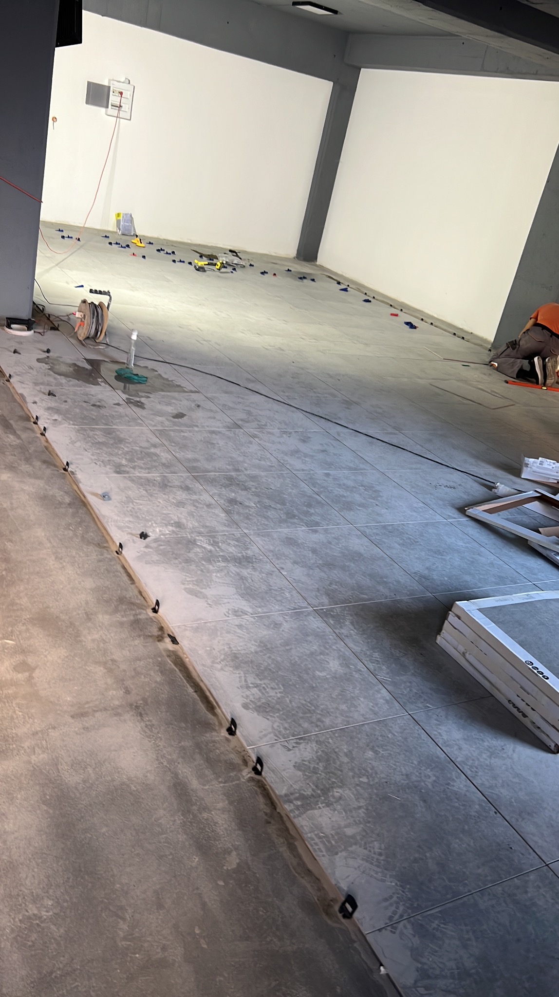 Układanie szarych płytek podłogowych w dużym pomieszczeniu, widoczne narzędzia i elementy poziomujące. Praca w toku, częściowo wykończona podłoga.