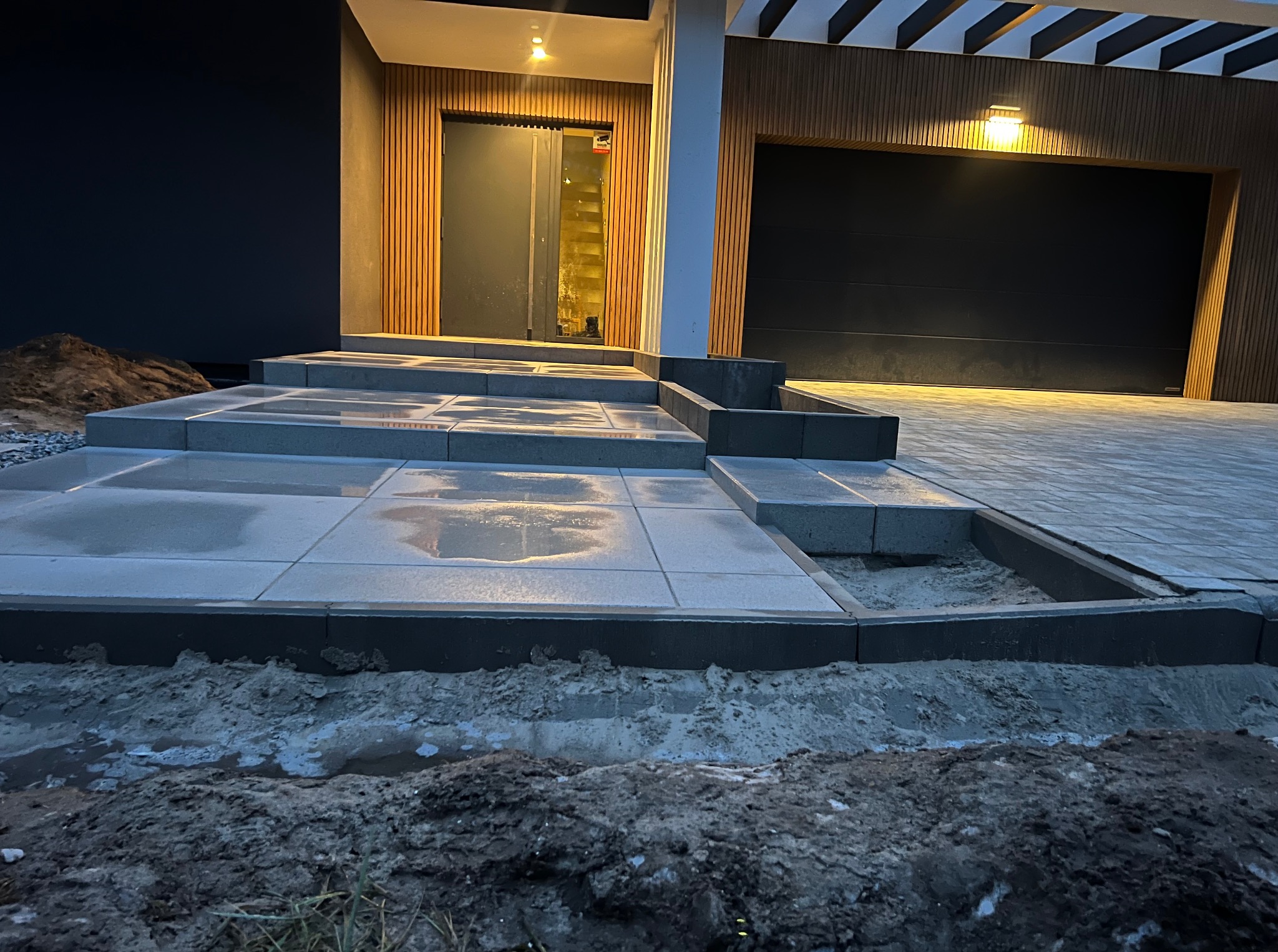 Nowoczesny dom z podjazdem wyłożonym szarą kostką brukową i schodami wejściowymi, oświetlony ciepłym światłem. Widoczne elementy wykończenia elewacji drewnem.