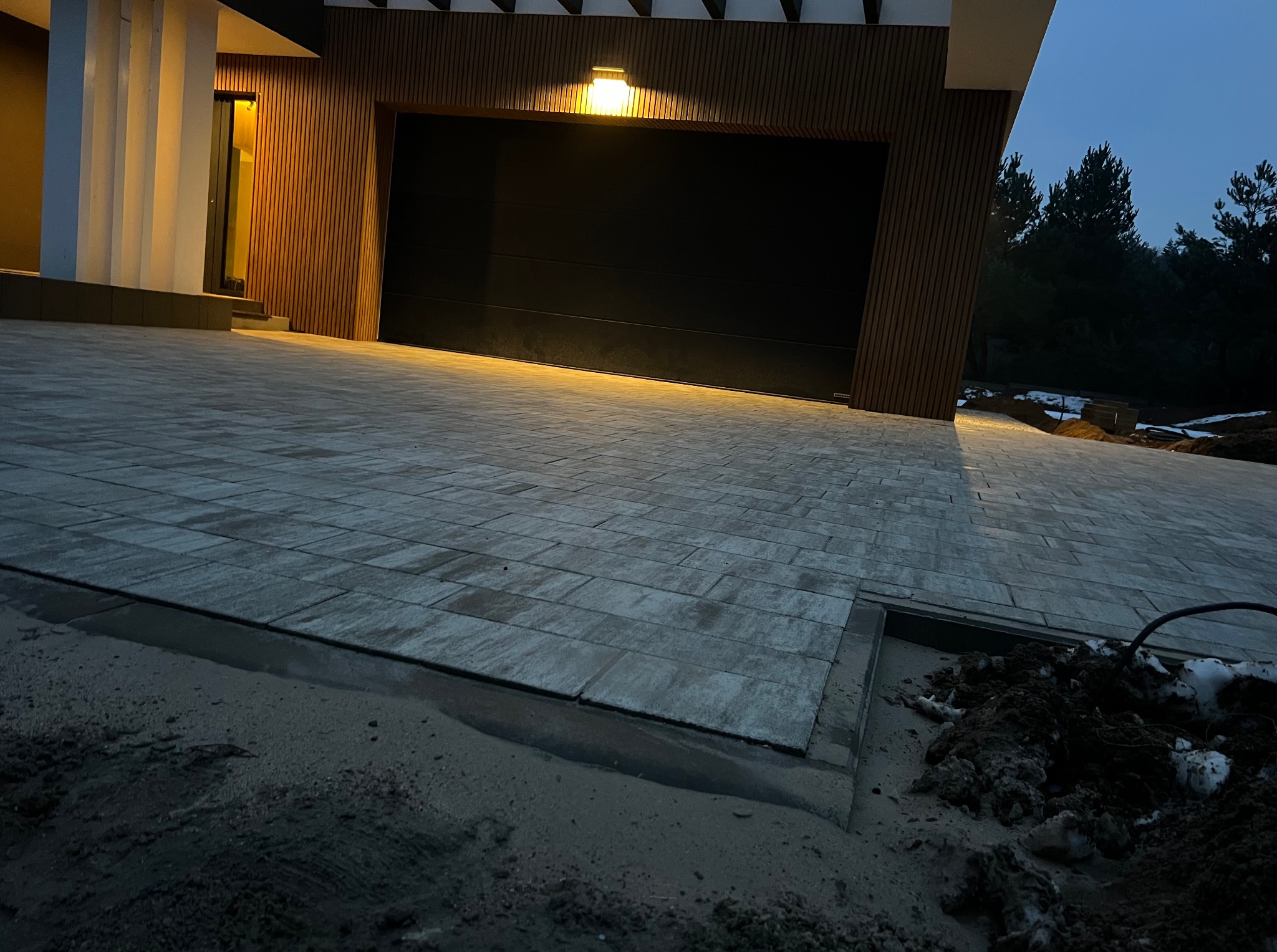 Nowoczesny podjazd z kostki brukowej w odcieniach szarości, oświetlony lampą nad garażem. Widoczne elementy elewacji budynku z drewnianymi akcentami i fragment ogrodu.