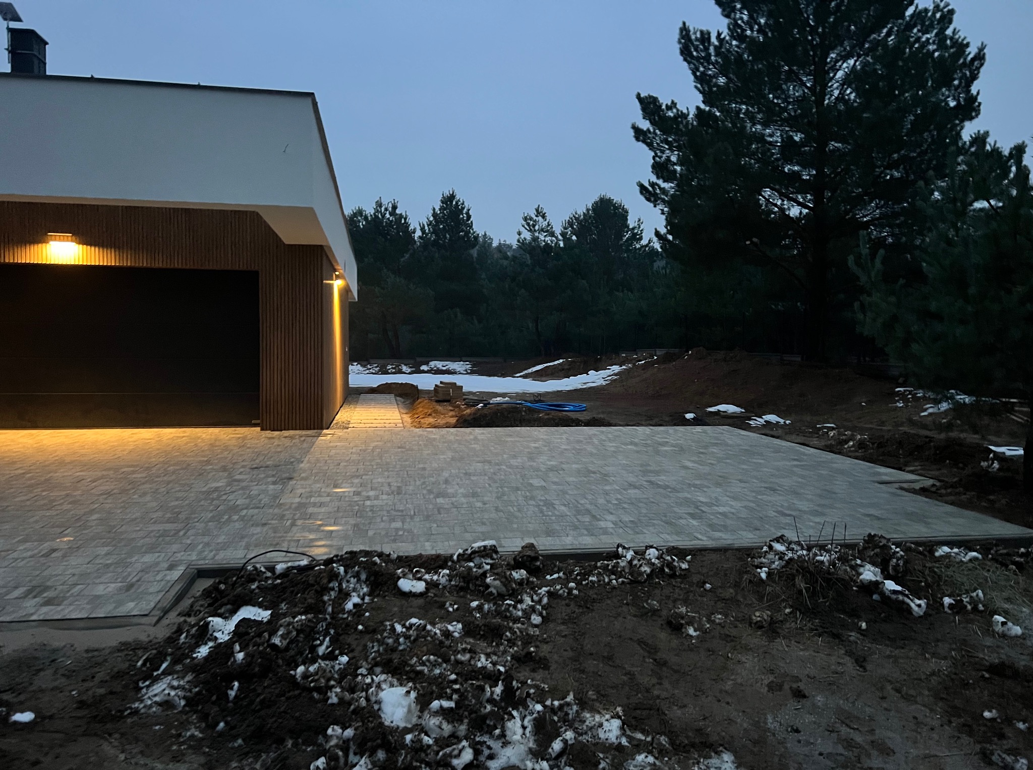 Ułożona kostka brukowa przed nowoczesnym domem z drewnianą elewacją i oświetlonym garażem, w tle las i resztki śniegu. Widok na podjazd w zimowej scenerii.