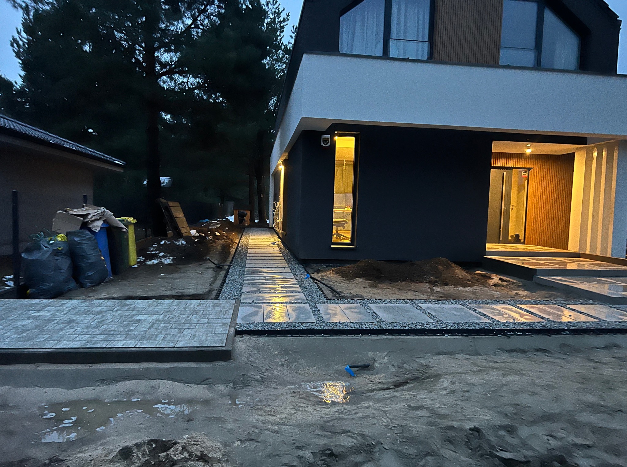 Nowoczesny dom z wybrukowaną ścieżką z płyt i kamienia. Ścieżka prowadzi do wejścia. Widoczne worki z materiałami budowlanymi. Oświetlenie domu włączone.