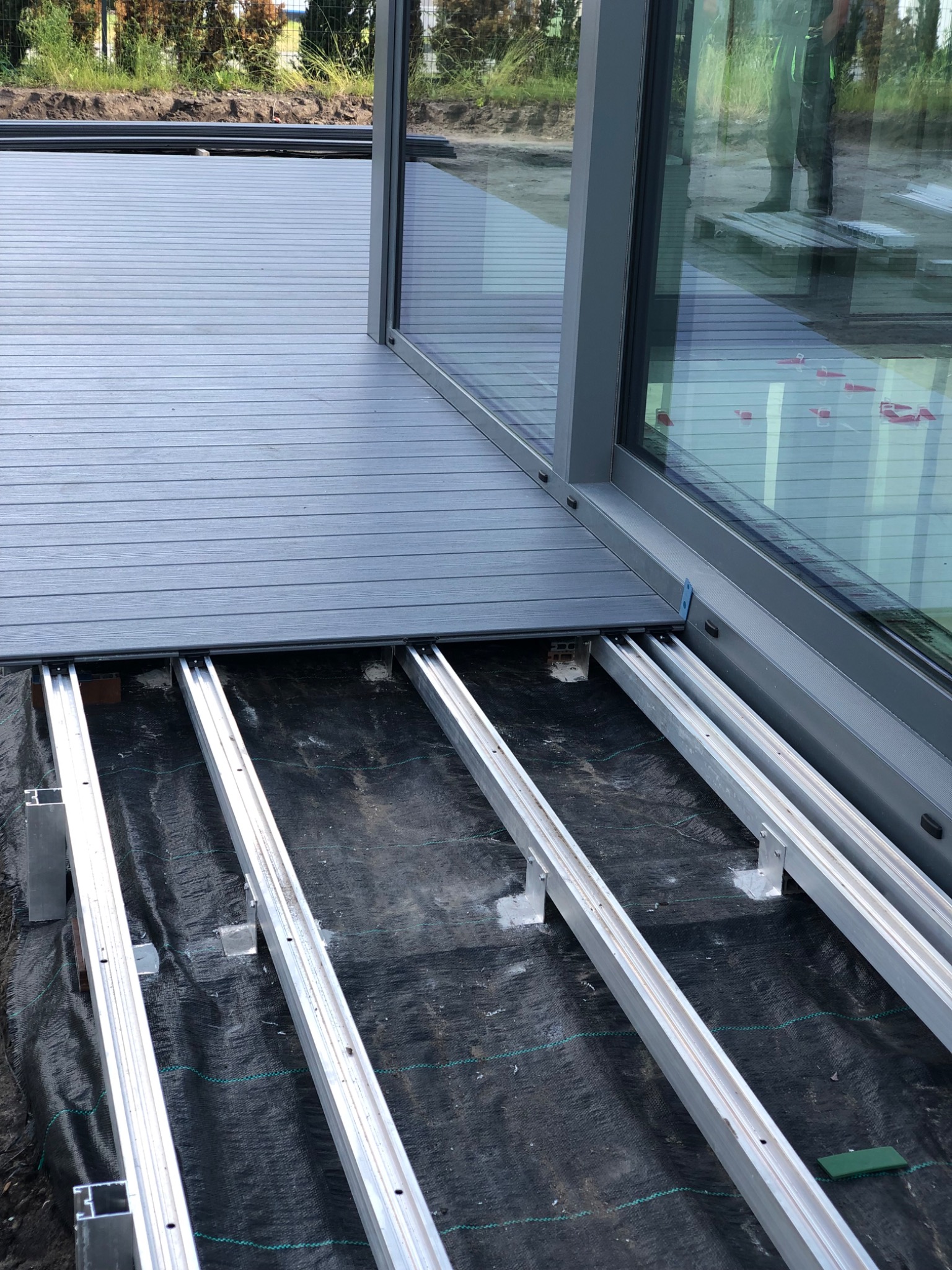 Montaż szarego tarasu: widoczne aluminiowe legary na czarnej membranie, częściowo ułożone deski. W tle okno tarasowe i zieleń.