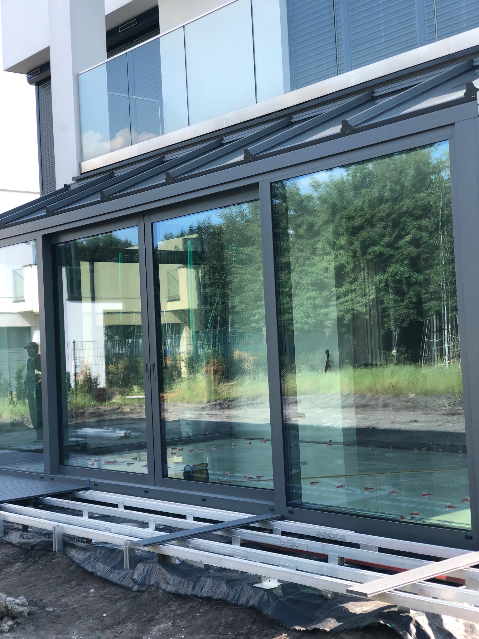 Szklana zabudowa tarasu w trakcie montażu, widoczne aluminiowe profile konstrukcyjne i częściowo ułożona podłoga. W tle zieleń i budynek mieszkalny.