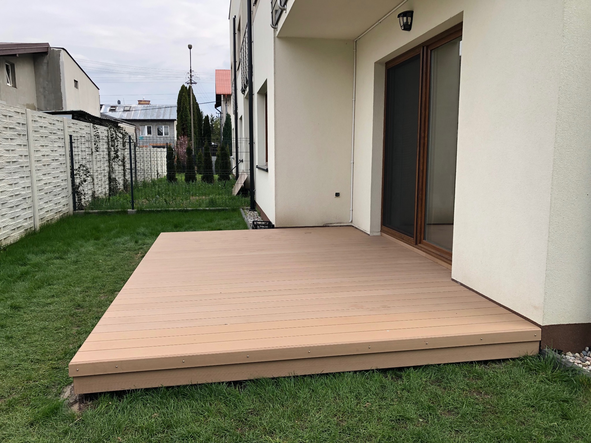 Jasny taras kompozytowy z desek ułożonych poziomo, przylegający do białego budynku z przesuwnymi drzwiami. Widok na trawnik i ogrodzenie w tle.