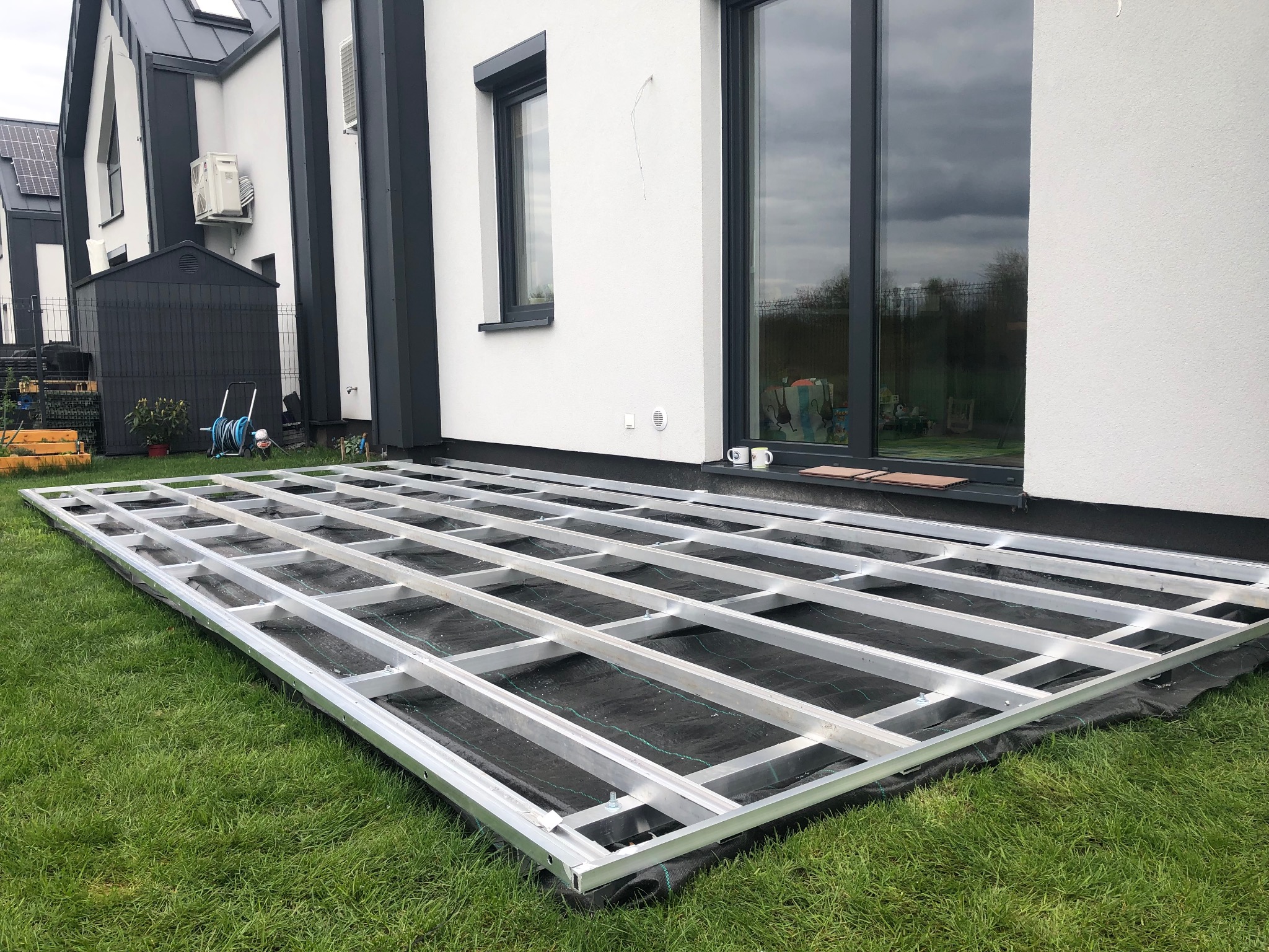 Metalowa konstrukcja pod taras na trawie, widok z góry. Elementy mocowane śrubami, czarna mata pod spodem. W tle dom z dużym oknem.