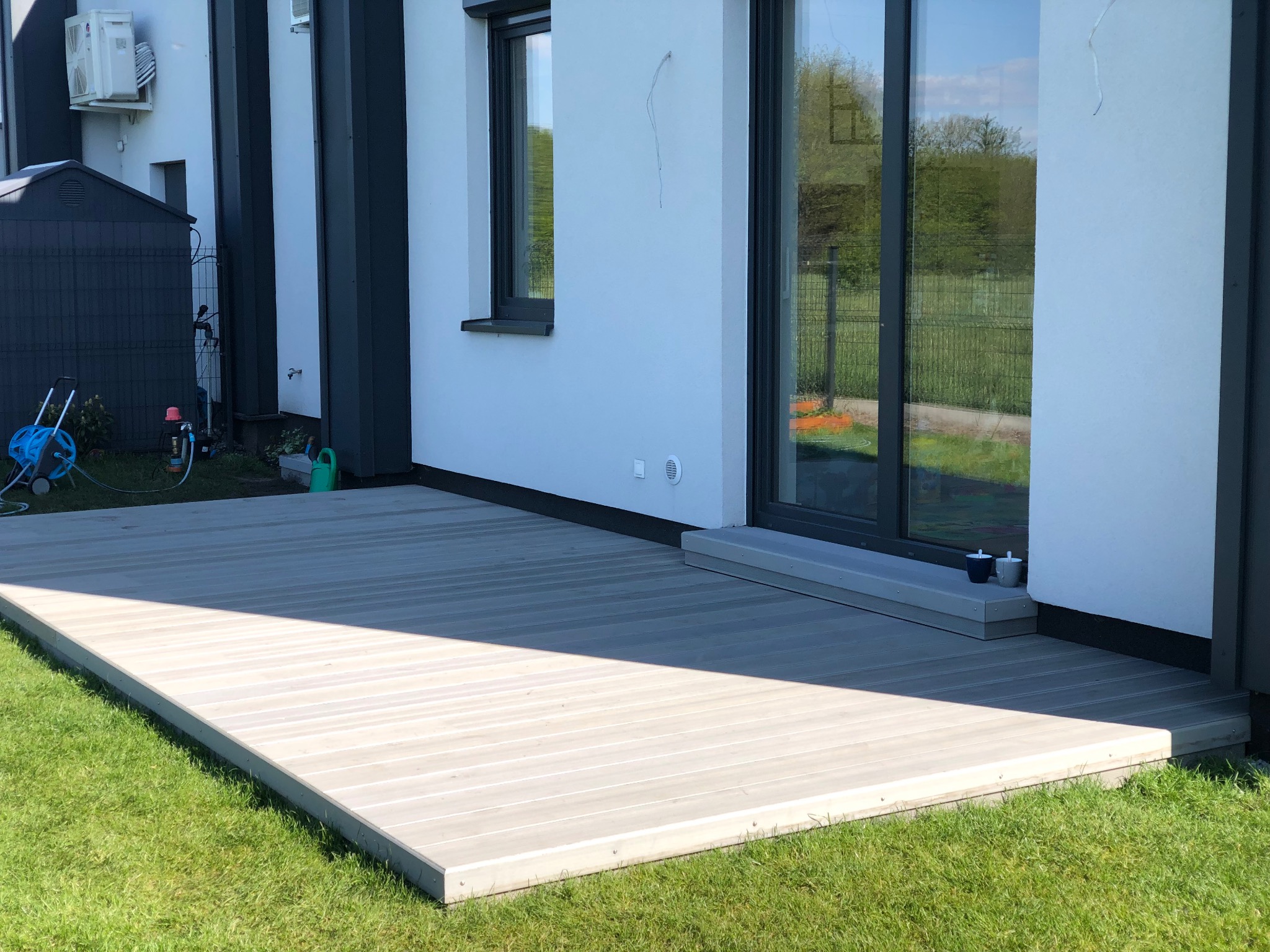 Jasny taras z desek kompozytowych przylegający do nowoczesnego domu z dużymi oknami. Widoczna zieleń trawnika i elementy architektury budynku.