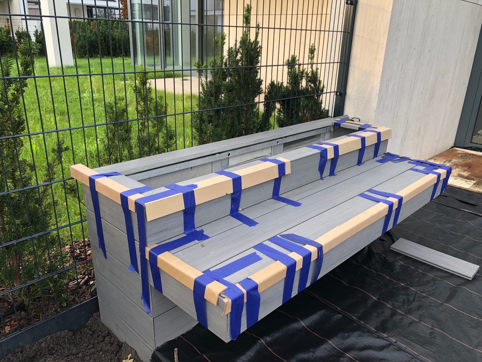 Montaż szarego tarasu kompozytowego z deskami zabezpieczonymi taśmą, widok z góry na fragment konstrukcji na czarnej macie, zieleń w tle za metalowym ogrodzeniem.