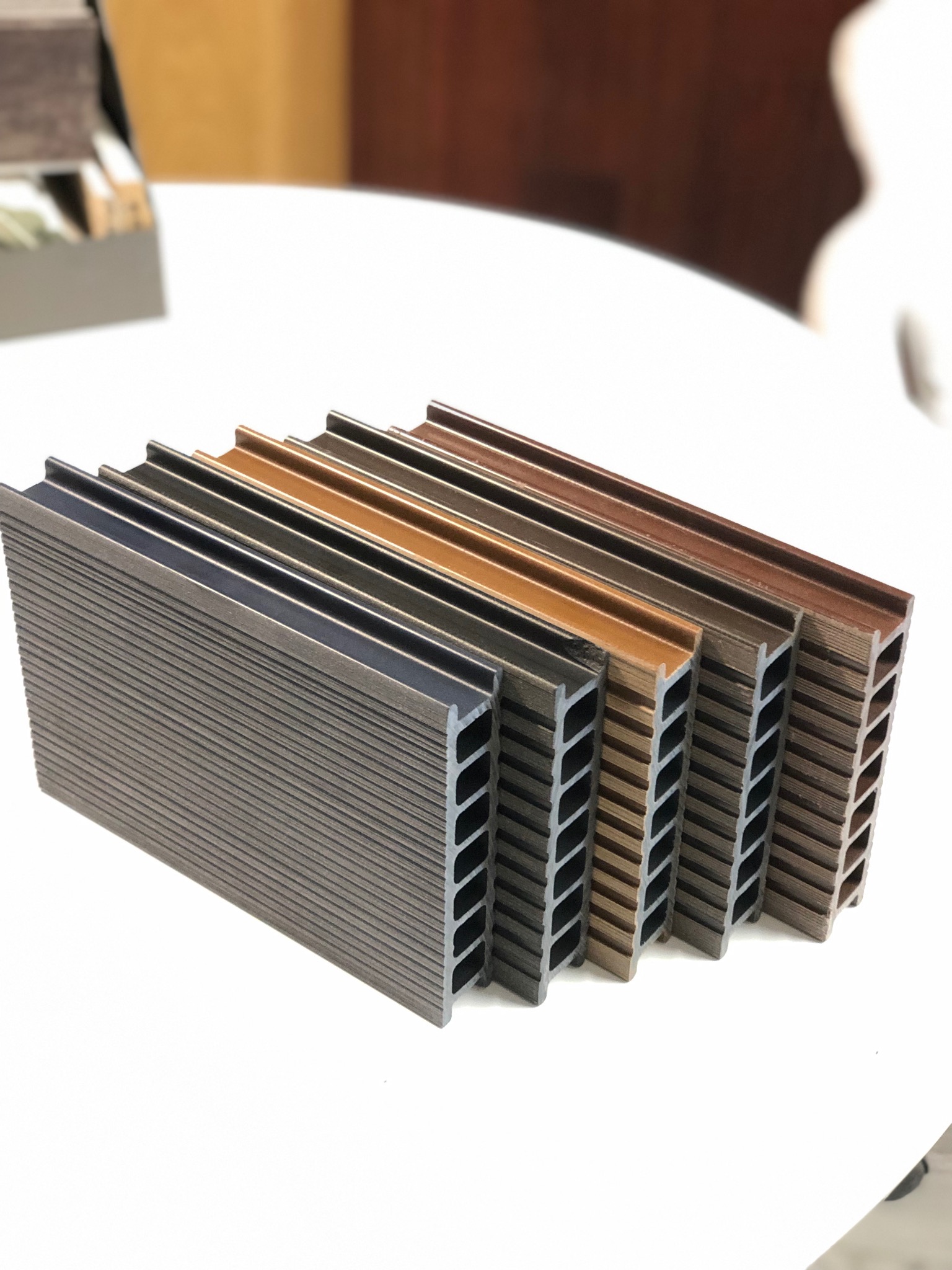 Przekrój poprzeczny próbek desek kompozytowych w różnych odcieniach brązu, ułożonych rzędami na białym tle. Widoczne żłobienia i wewnętrzna struktura.