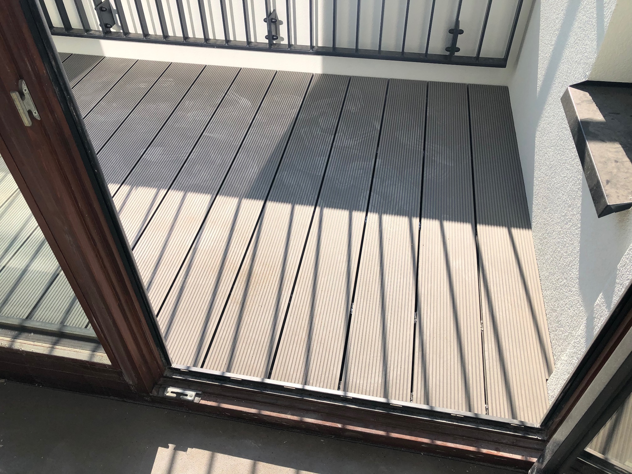 Jasny balkon z szarą podłogą kompozytową, widok z wnętrza przez otwarte drzwi balkonowe. Cień barierki na podłodze. Minimalistyczny styl.