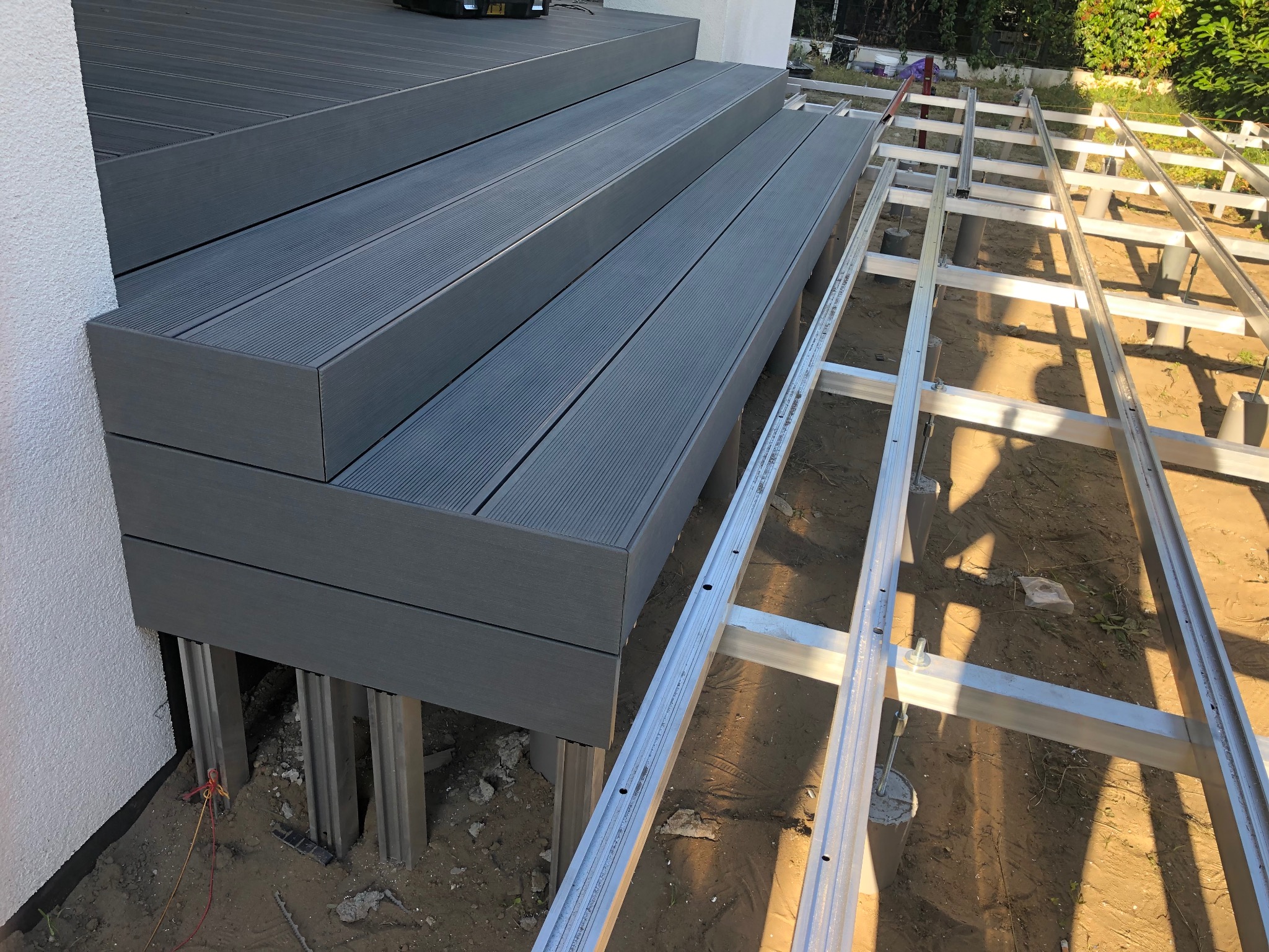 Montaż tarasu kompozytowego: widoczne schody z desek i aluminiowa konstrukcja nośna na betonowych słupach. Częściowo ukończony projekt tarasu.