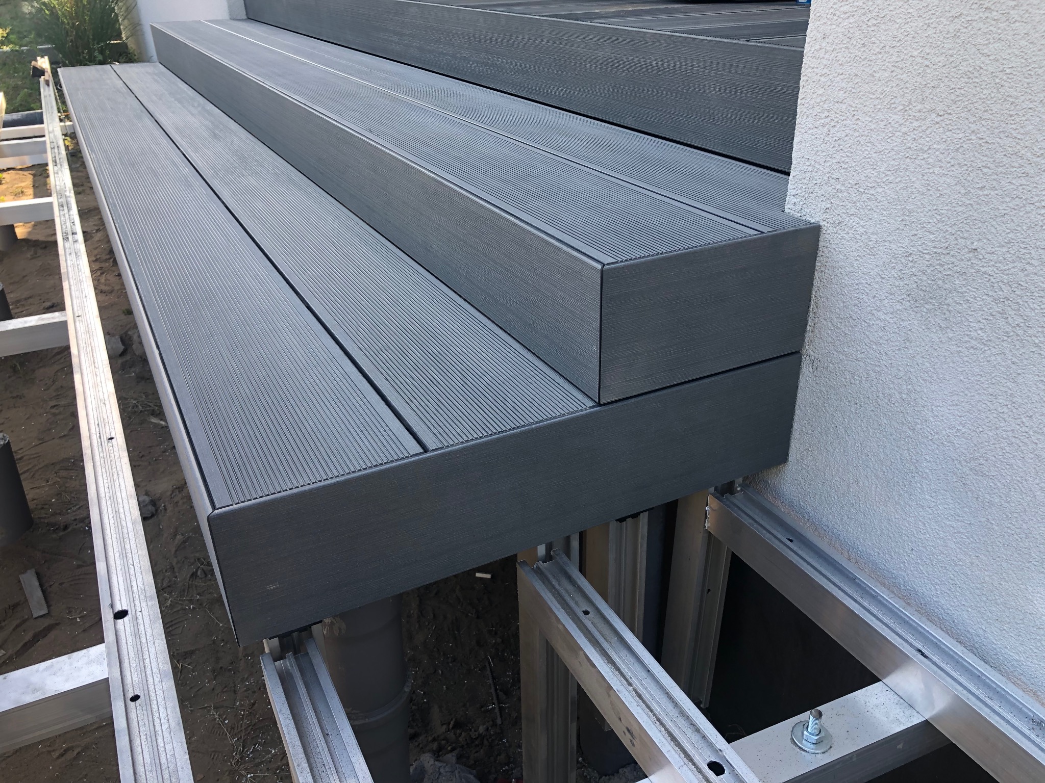 Nowoczesny taras z deski kompozytowej w odcieniach szarości, z widoczną aluminiową konstrukcją nośną i fragmentem elewacji budynku. Detal wykończenia schodów.